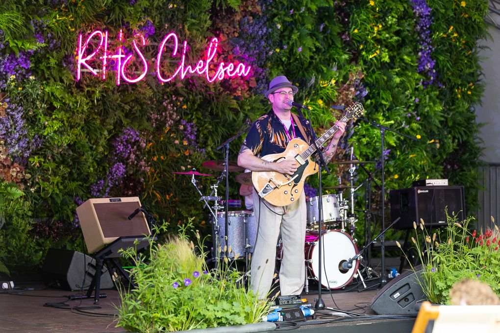 A musician performs on stage with a guitar beneath pink neon signage and lush greenery backdrop.