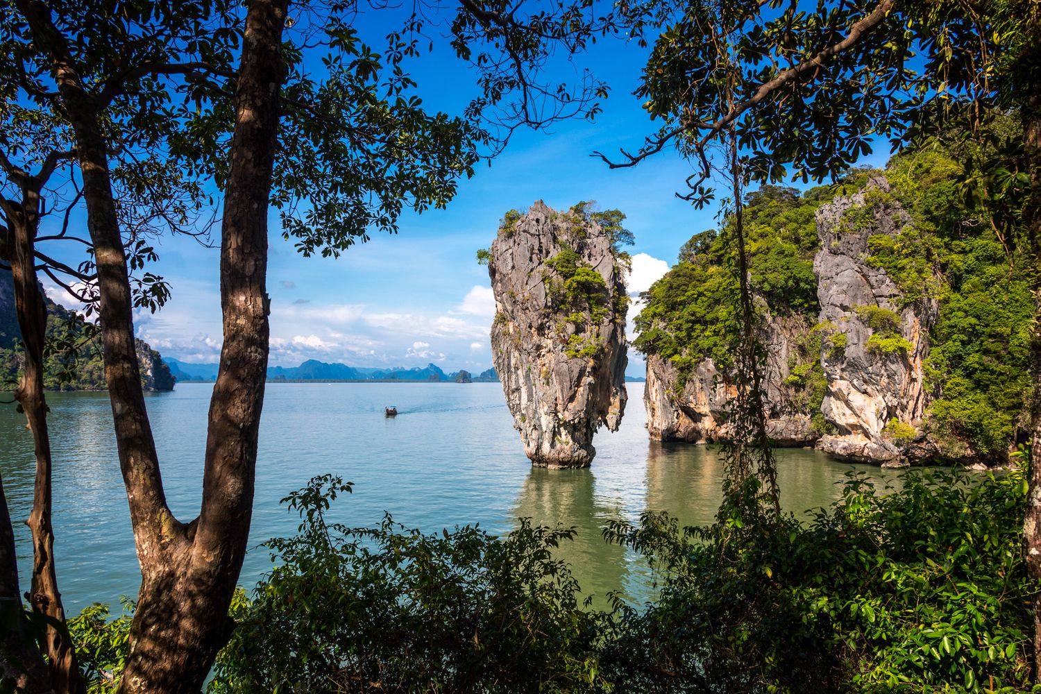 Dramatic limestone karst formation rises from turquoise waters in Phang Nga Bay, Thailand, framed by tropical trees.