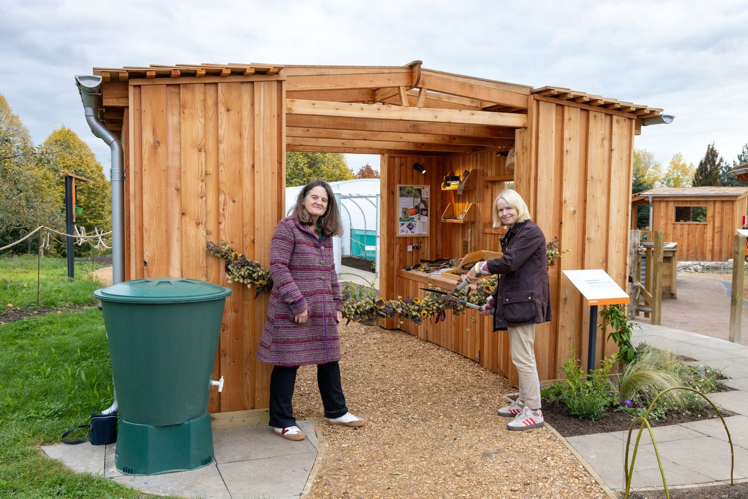 Two people stand outside a wooden garden shed with a green rain barrel in a community garden setting.