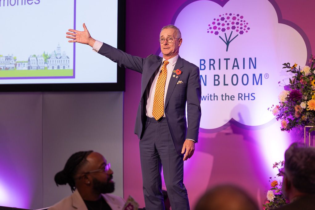 A speaker presents on stage at a Britain in Bloom event with purple lighting and presentation screens visible.