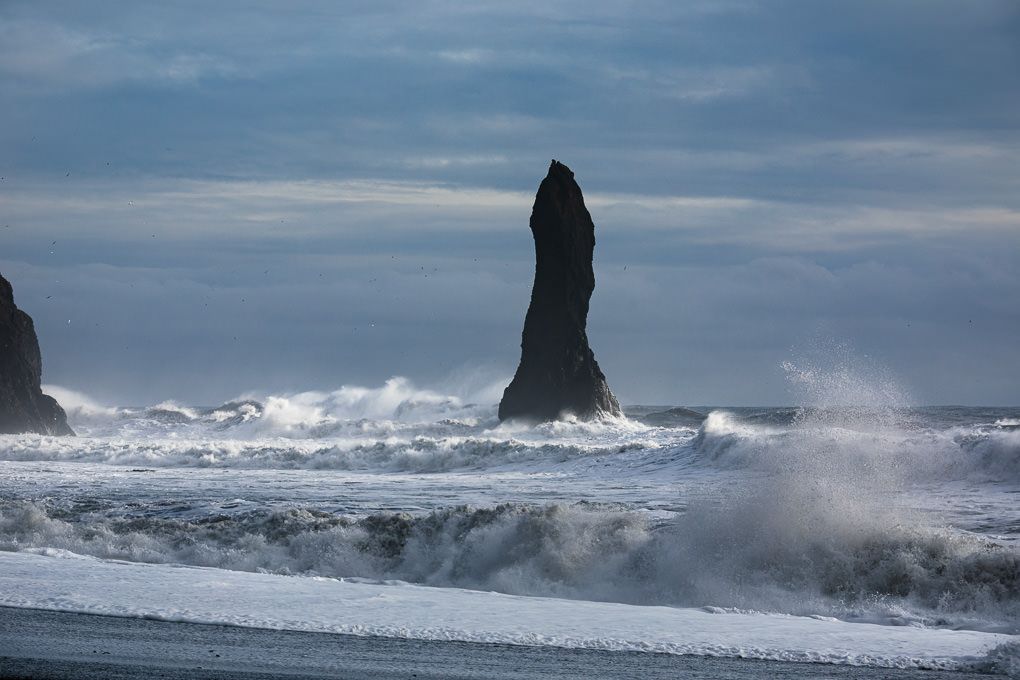 Dramatic waves crash against a towering sea stack on a black sand beach in Iceland under moody skies.