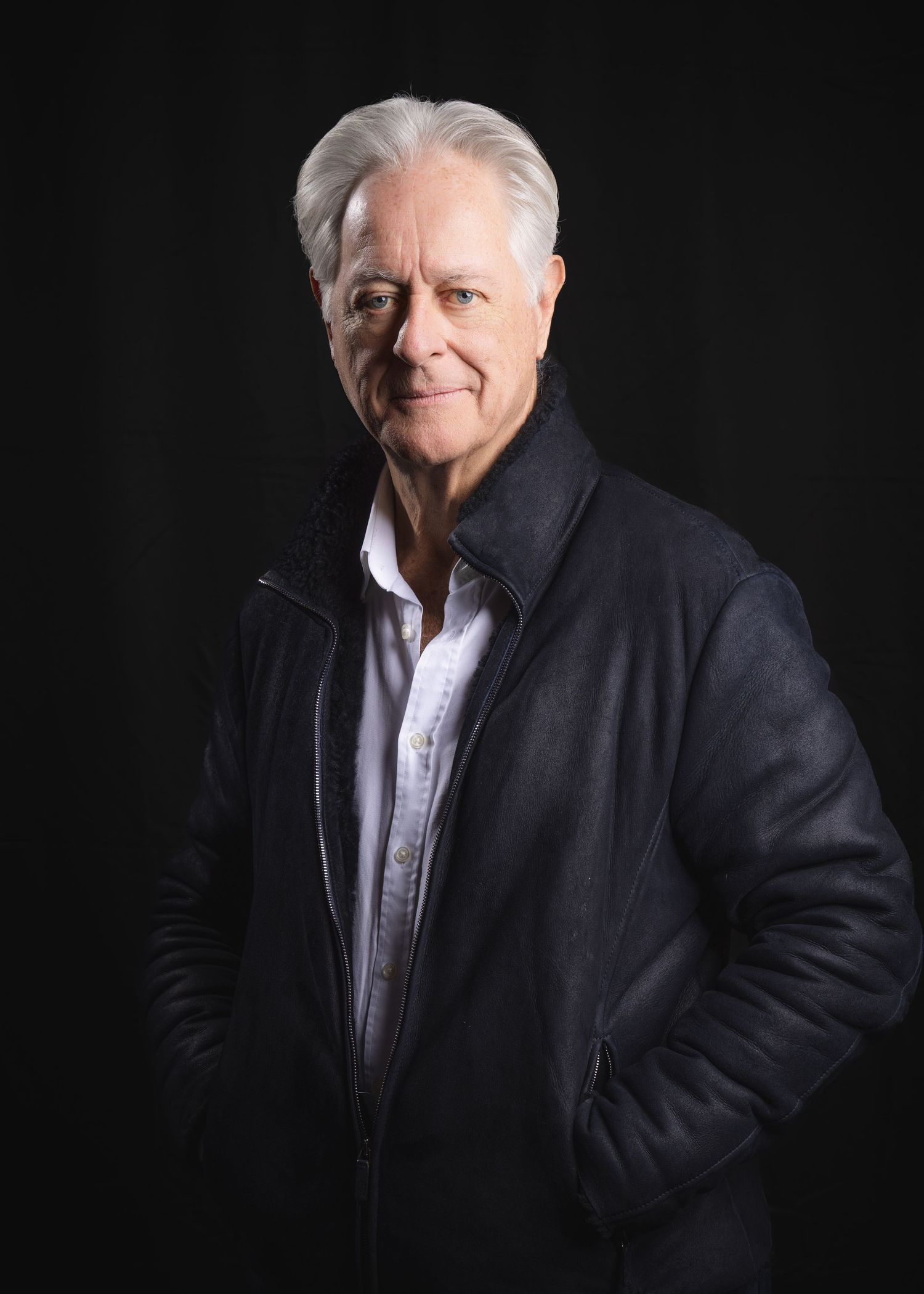 Confident older man with silver hair wearing a dark leather jacket poses against a black background.