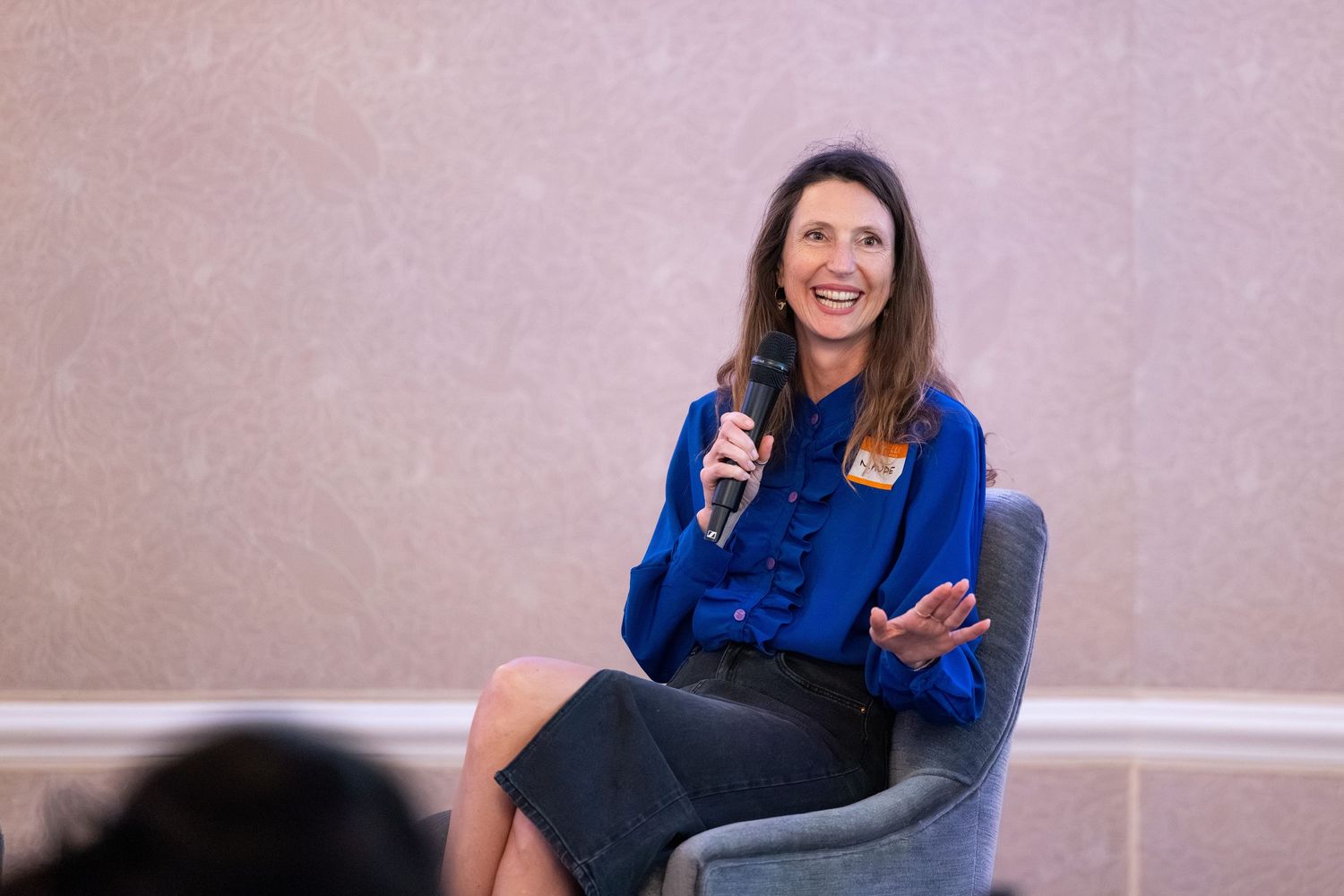 Woman in blue blouse speaks with microphone while seated in gray chair at professional event.