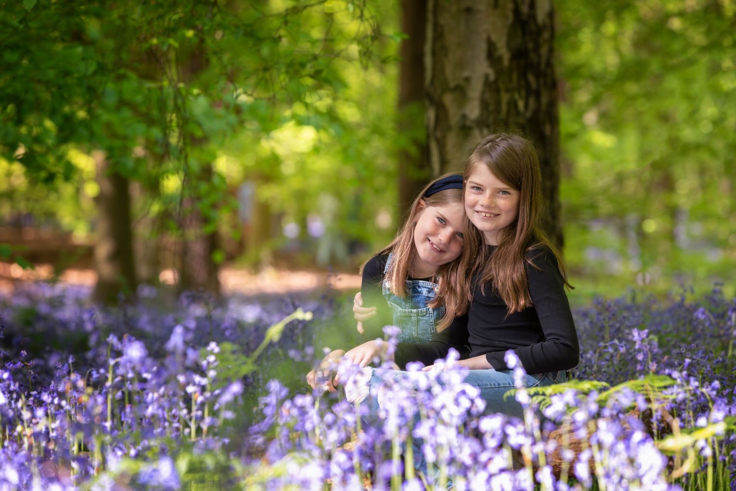 Two girls smiling among purple bluebells in a sunlit woodland forest setting.