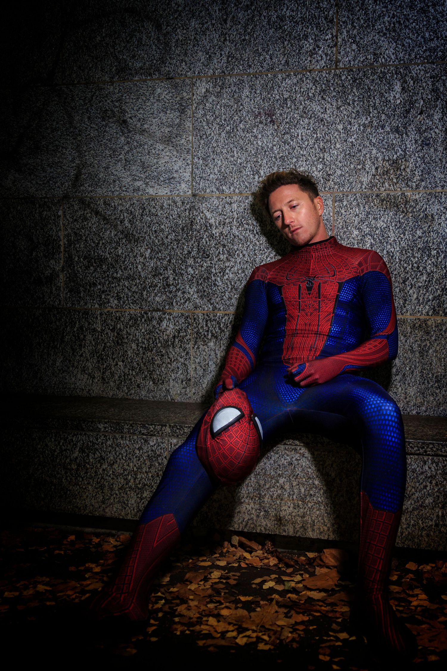 Cosplay portrait of Spider-Man character sitting against a stone wall, holding mask with dramatic lighting and autumn leaves.