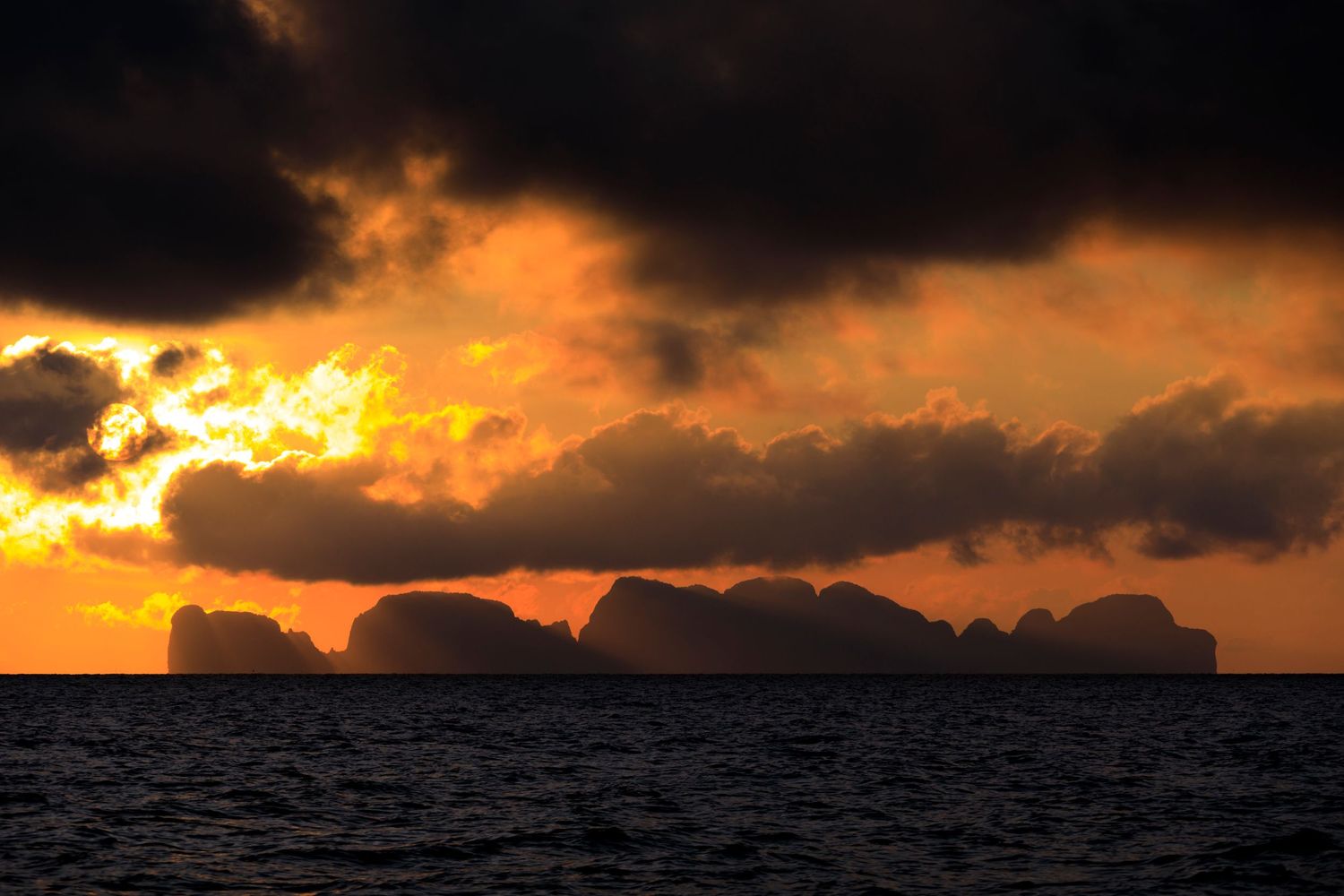 Dramatic golden sunset illuminates dark storm clouds over silhouetted rocky islands on a moody ocean horizon.