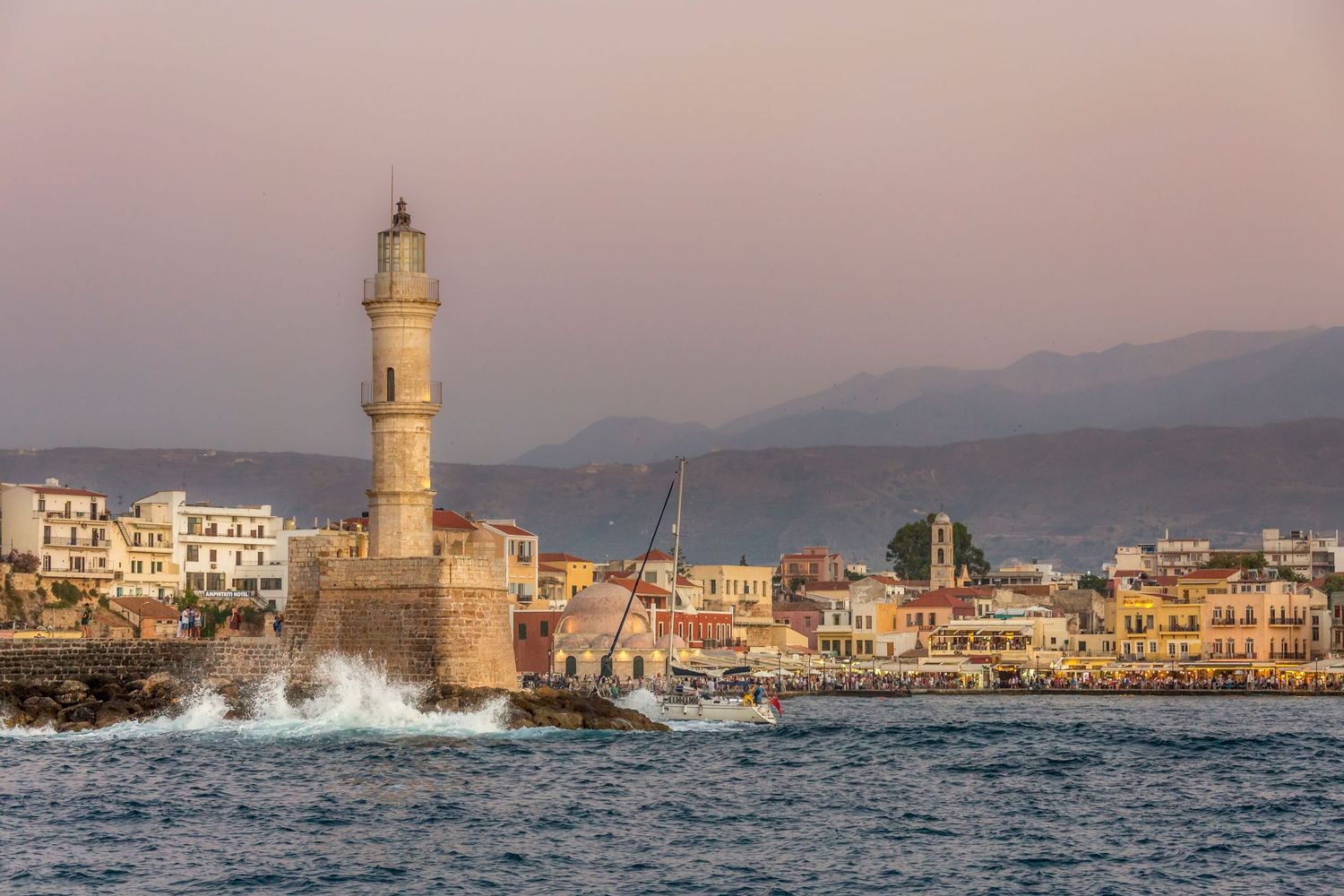 Historic Venetian lighthouse at Chania harbour, Crete, Greece, at sunset with crashing waves and colorful waterfront buildings.
