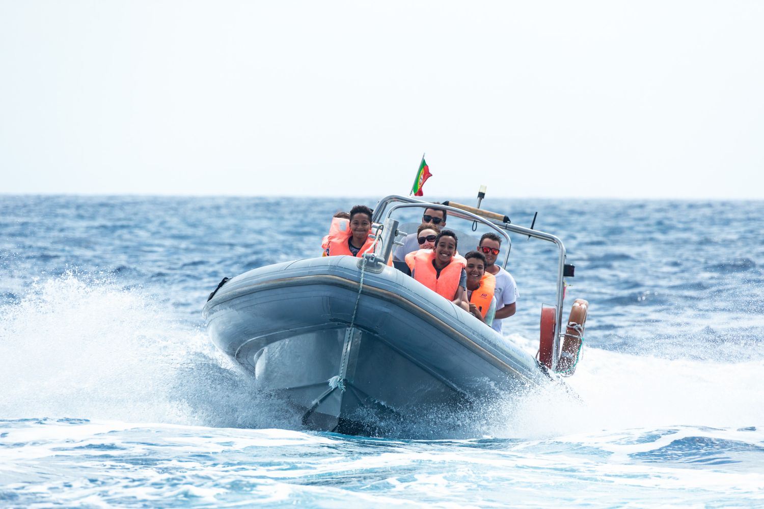 A rigid inflatable rescue boat speeds across blue ocean waters with passengers in orange life vests.