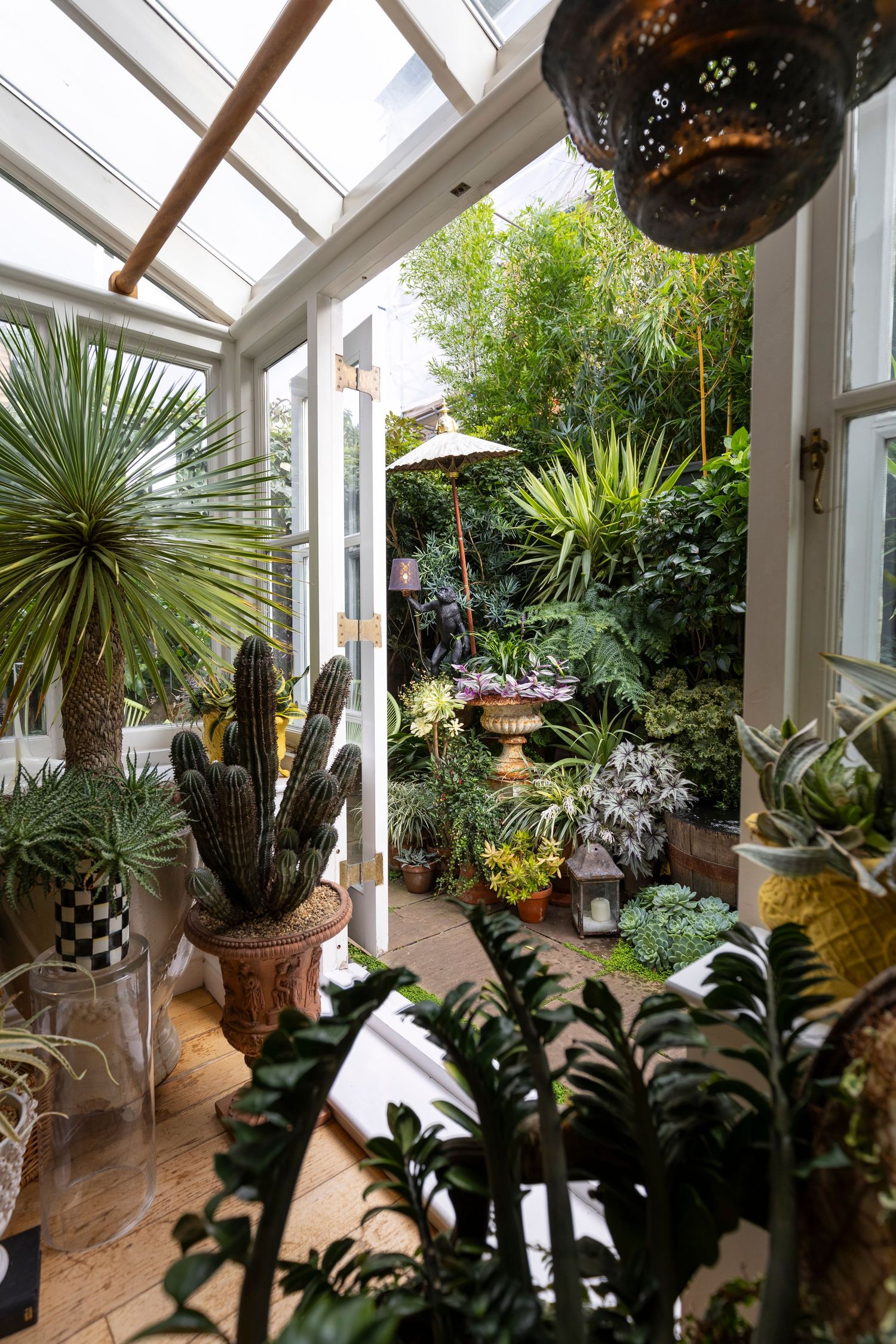 Lush conservatory filled with cacti, tropical plants, and succulents under a glass roof, opening to a verdant garden patio.