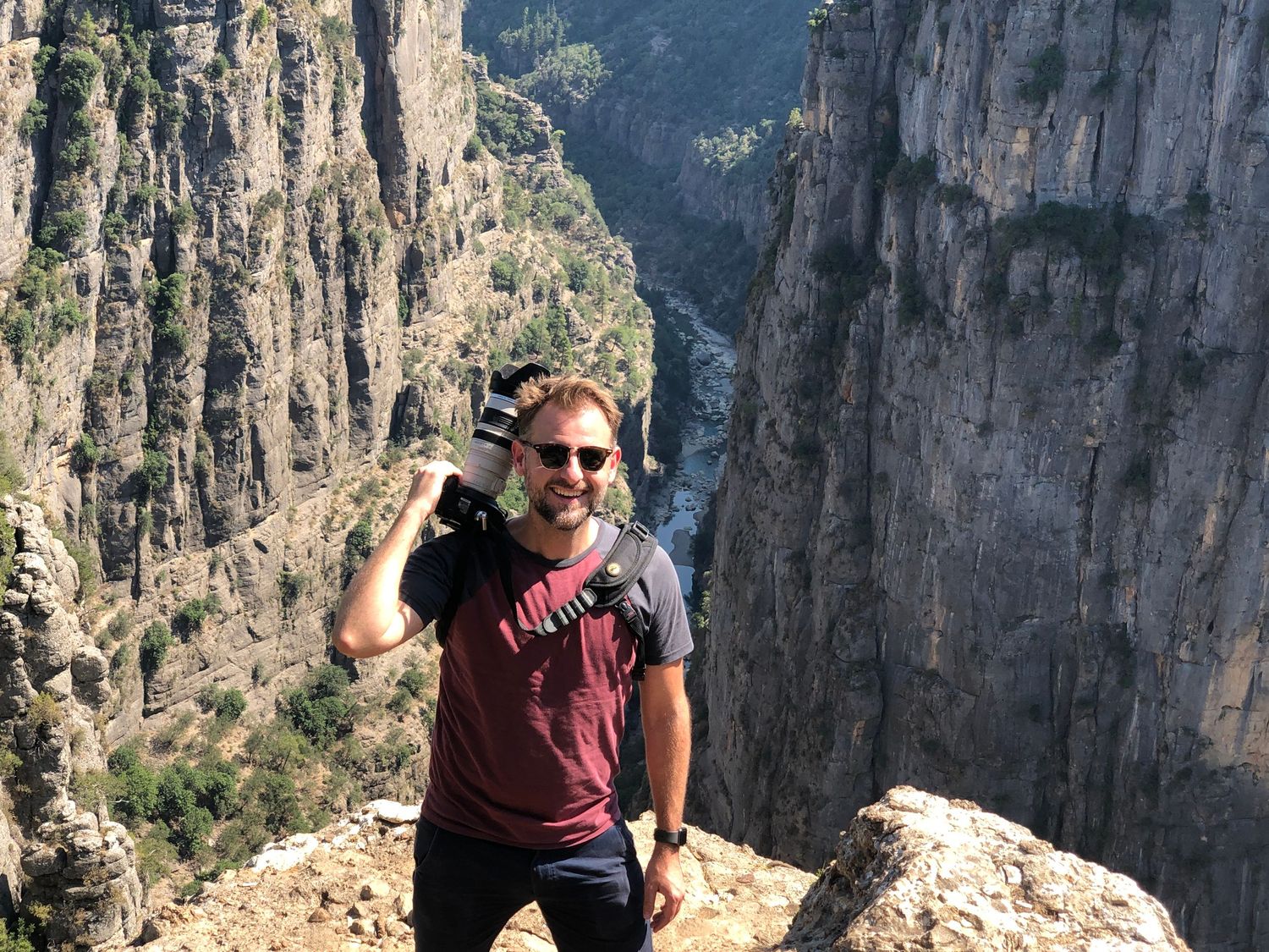 Man holding camera at canyon overlook with dramatic rocky cliffs and deep gorge in background.