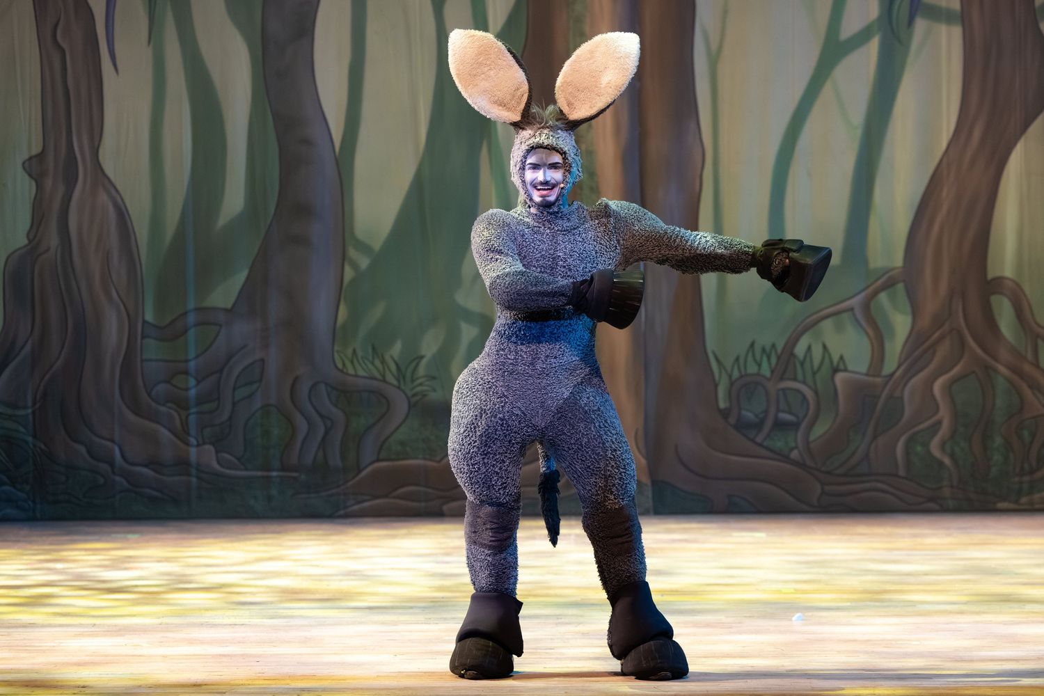 Donkey character in costume strikes a dramatic pose on a forest-themed stage set during Shrek The Musical in Wellingborough