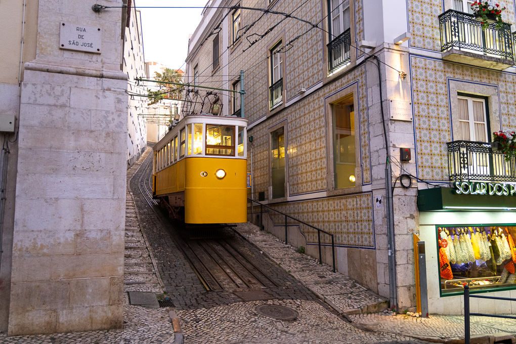Historic yellow funicular tram car climbs steep rails through narrow streets of Lisbon's Bairro Alto district.