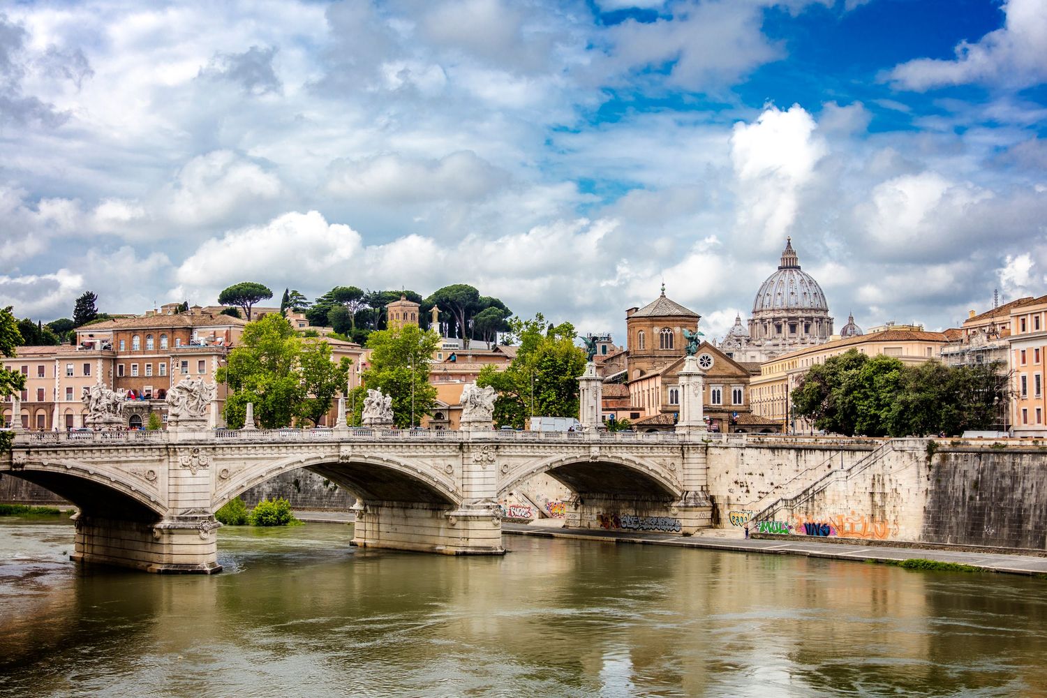 A stone bridge spans the Tiber River with St. Peters Basilica dome visible on the skyline in Rome.