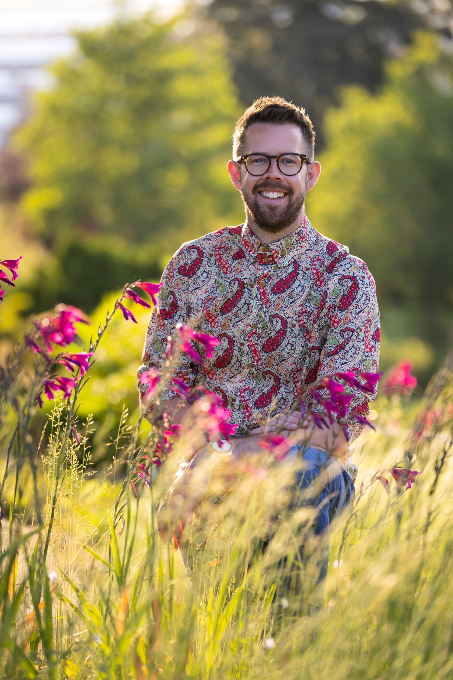 Smiling bearded man with glasses wearing a colorful paisley shirt standing in a sunny wildflower garden.