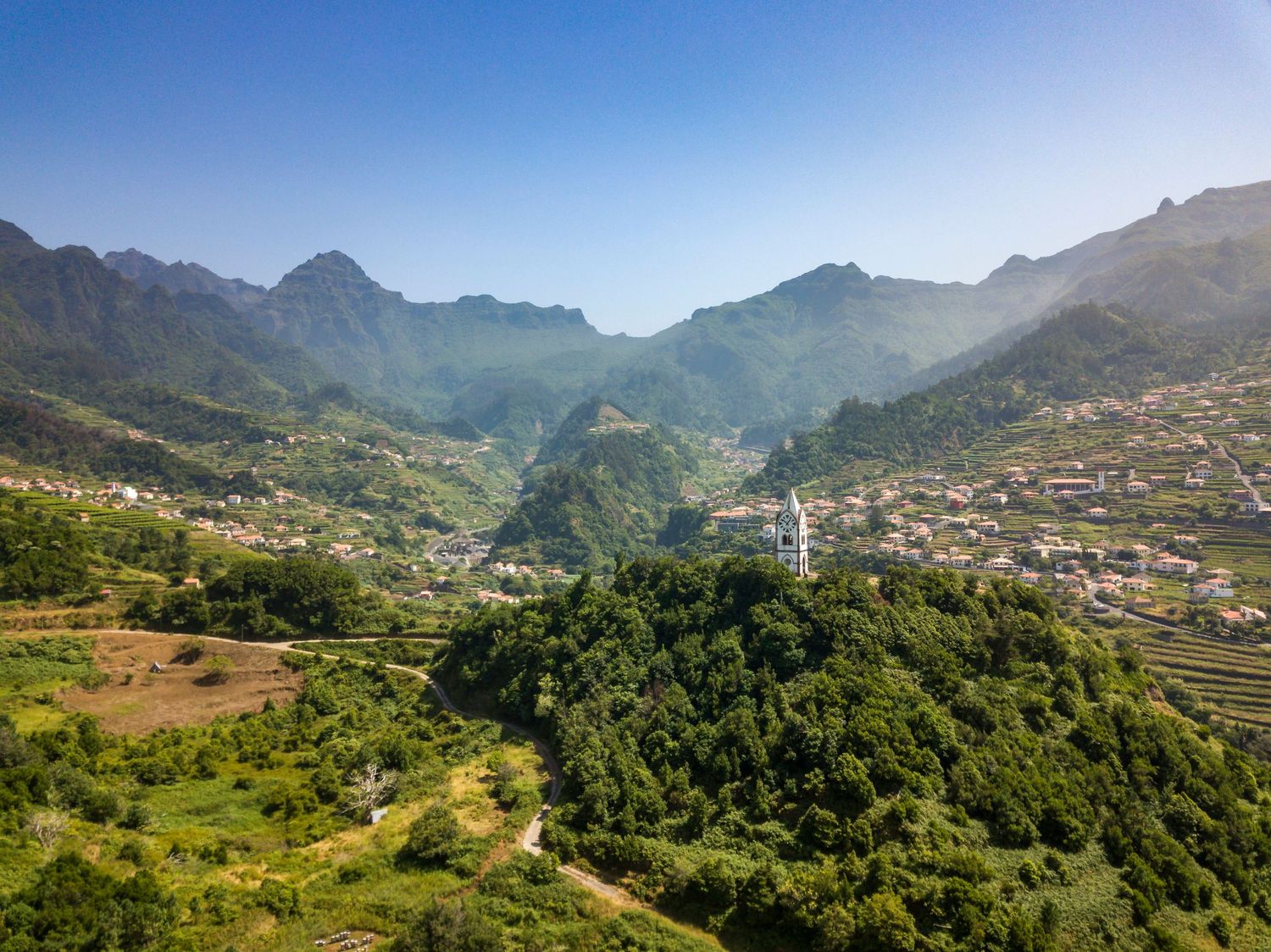 Aerial view of a lush green mountain valley with a small village nestled among misty peaks under a clear blue sky.