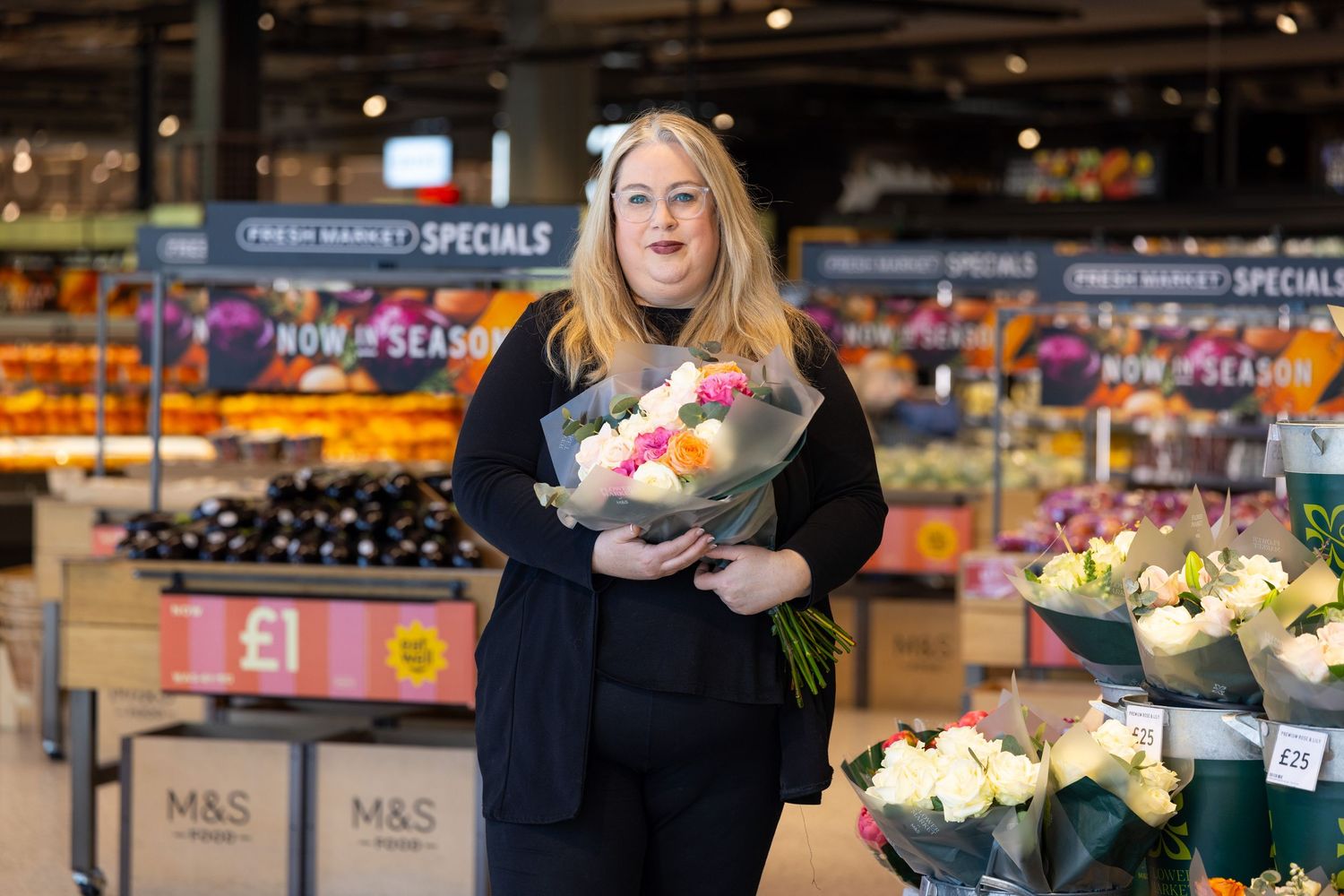 Woman holding a bouquet of flowers in an M&S Fresh Market grocery store with colorful produce displays.