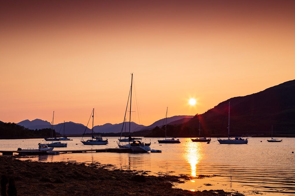 Silhouettes of sailboats anchored in Glencoe, Scotland, during a vibrant orange sunset with mountains in the background.