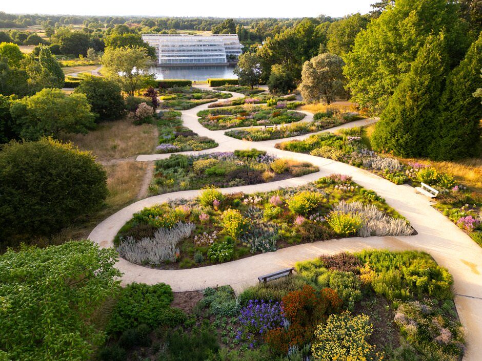 Aerial view of winding garden paths through colorful flower beds with a white building in the background.