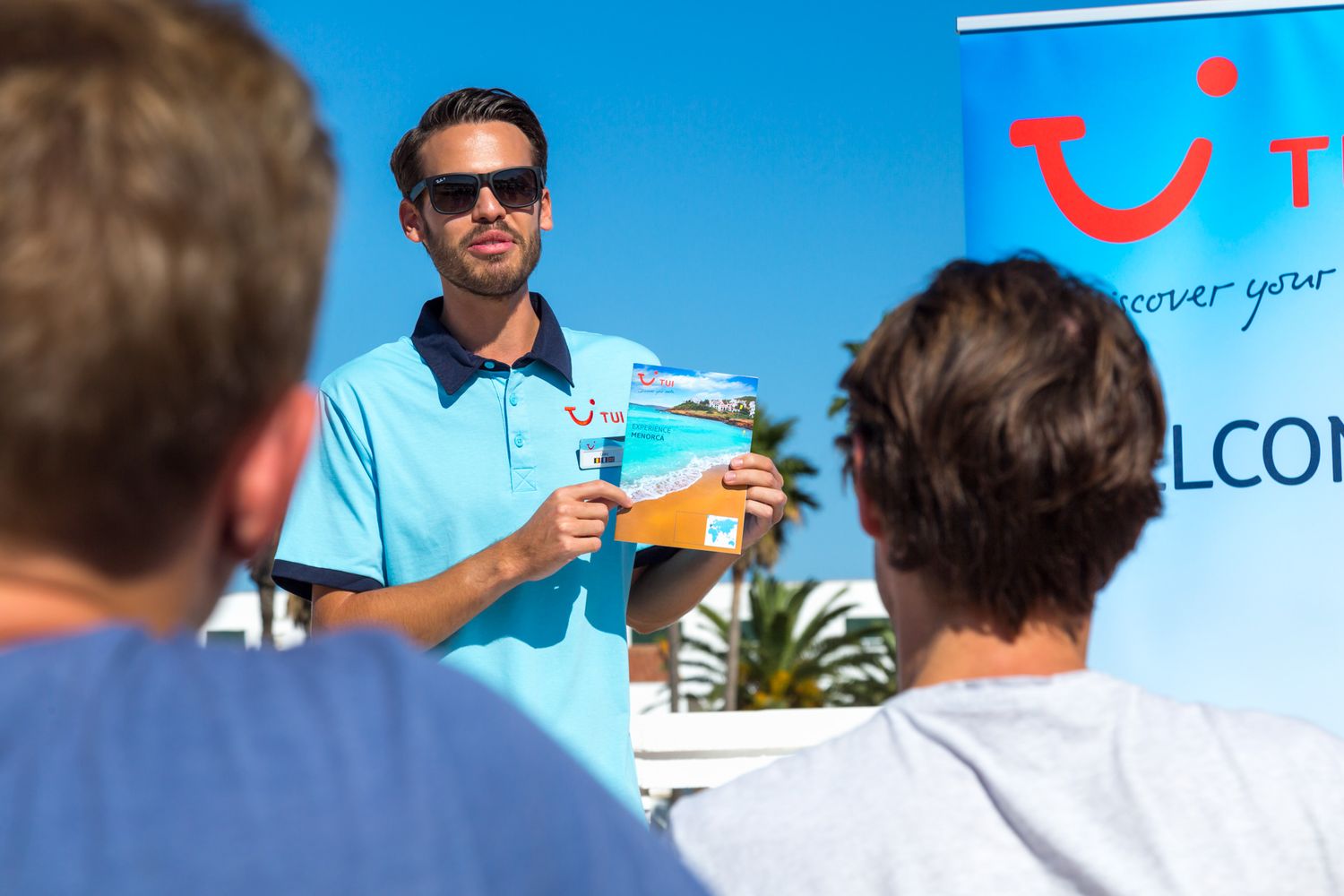 A TUI rep in blue polo shirt speaking to tourists at outdoor gathering with TUI Smile logo visible in background.