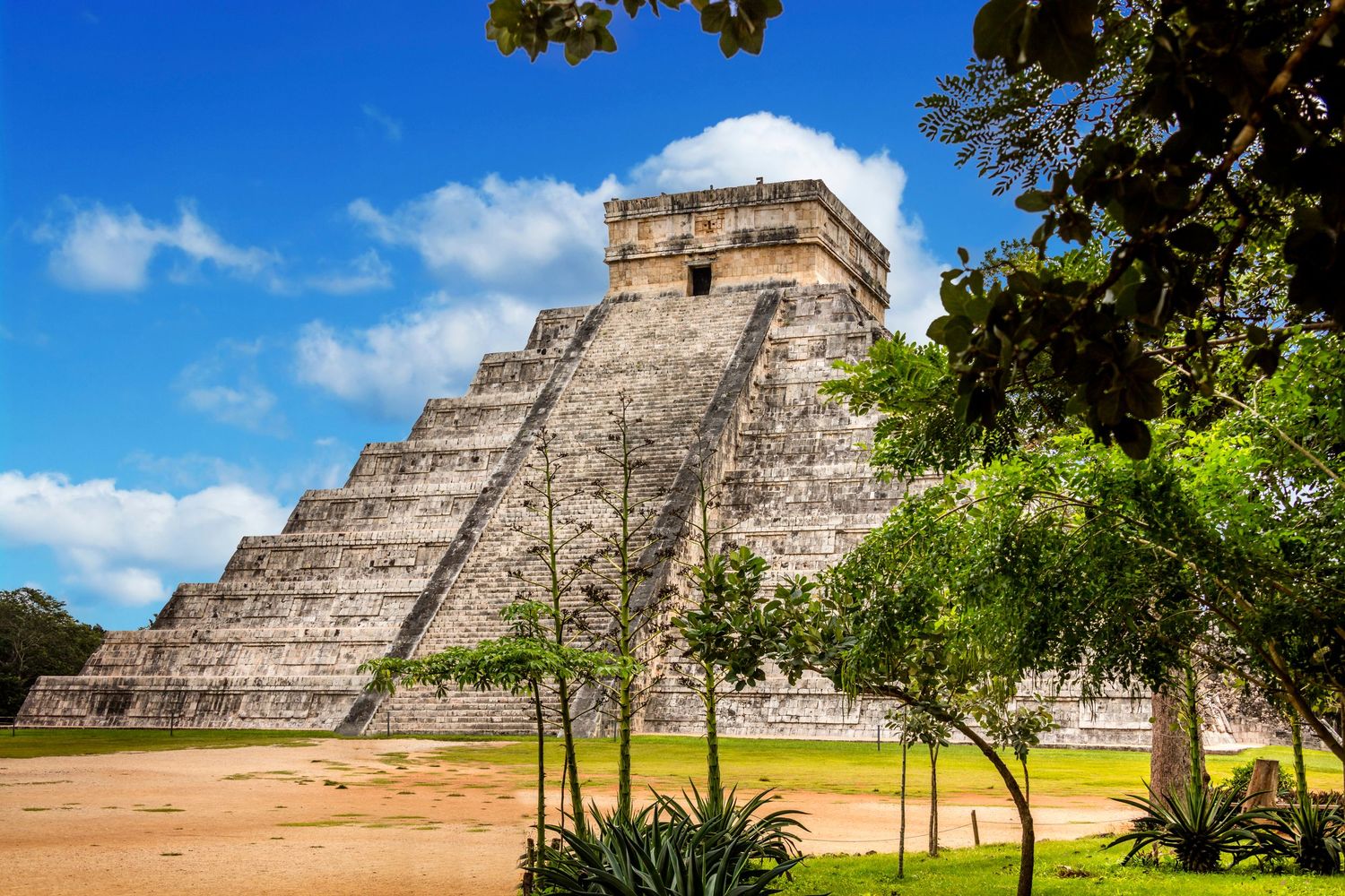 Ancient Mayan pyramid of Chichen Itza rising above tropical trees under a bright blue sky in Mexico.