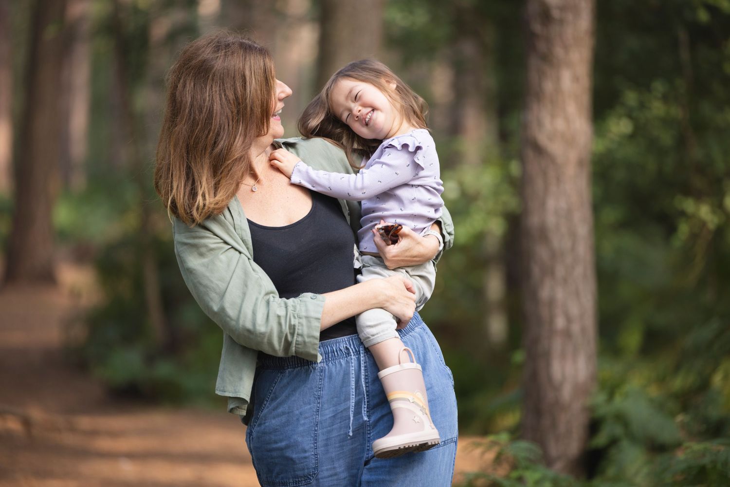 A mother holds her laughing toddler in a sunlit forest, both wearing casual outfits, sharing a joyful embrace.