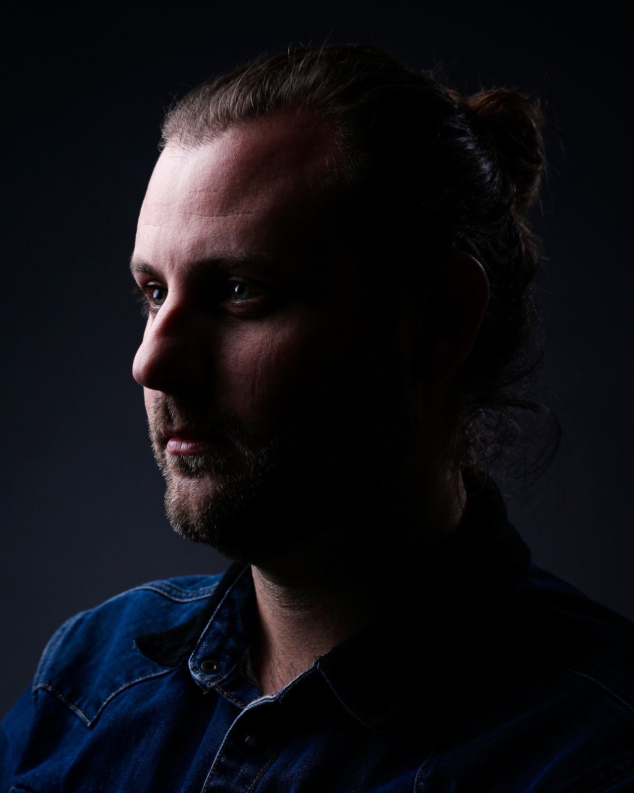 Dramatic low-key portrait of a bearded man with hair in a bun, wearing a denim shirt against a dark background.
