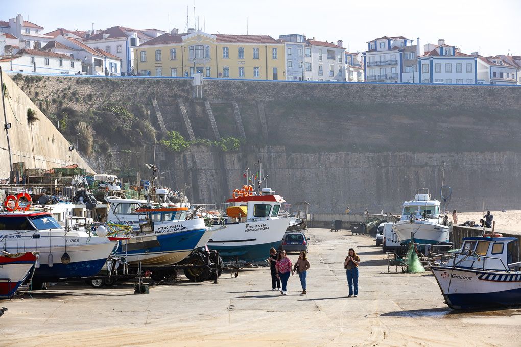 Small fishing boats line the beach below steep coastal cliffs with colorful buildings perched along the top in a seaside town.