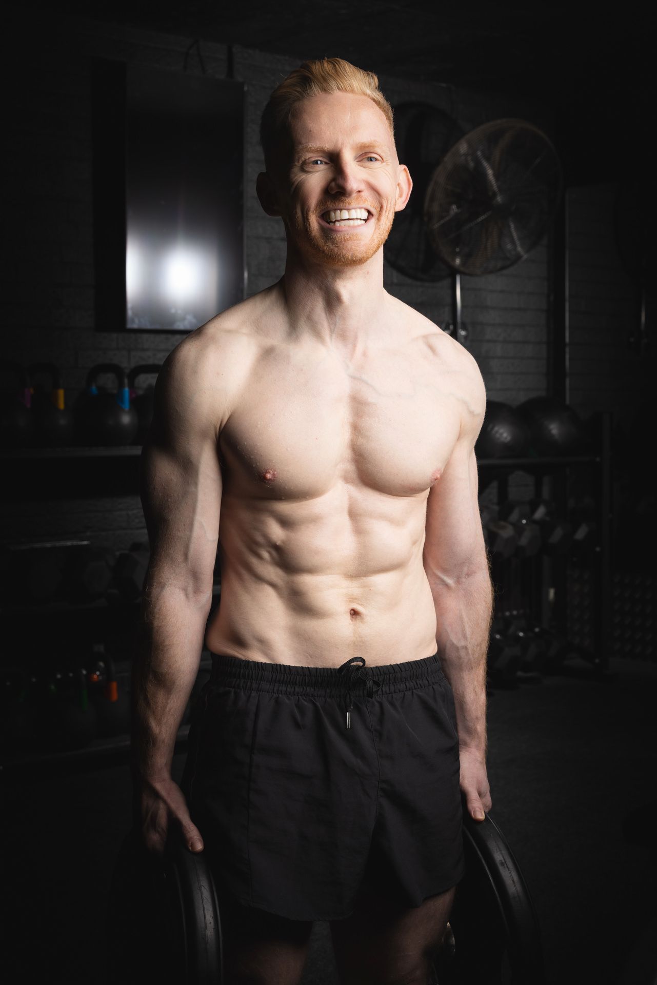 Fitness photoshoot. Athletic male with muscular physique posing shirtless in a dark Hertfordshire gym.
