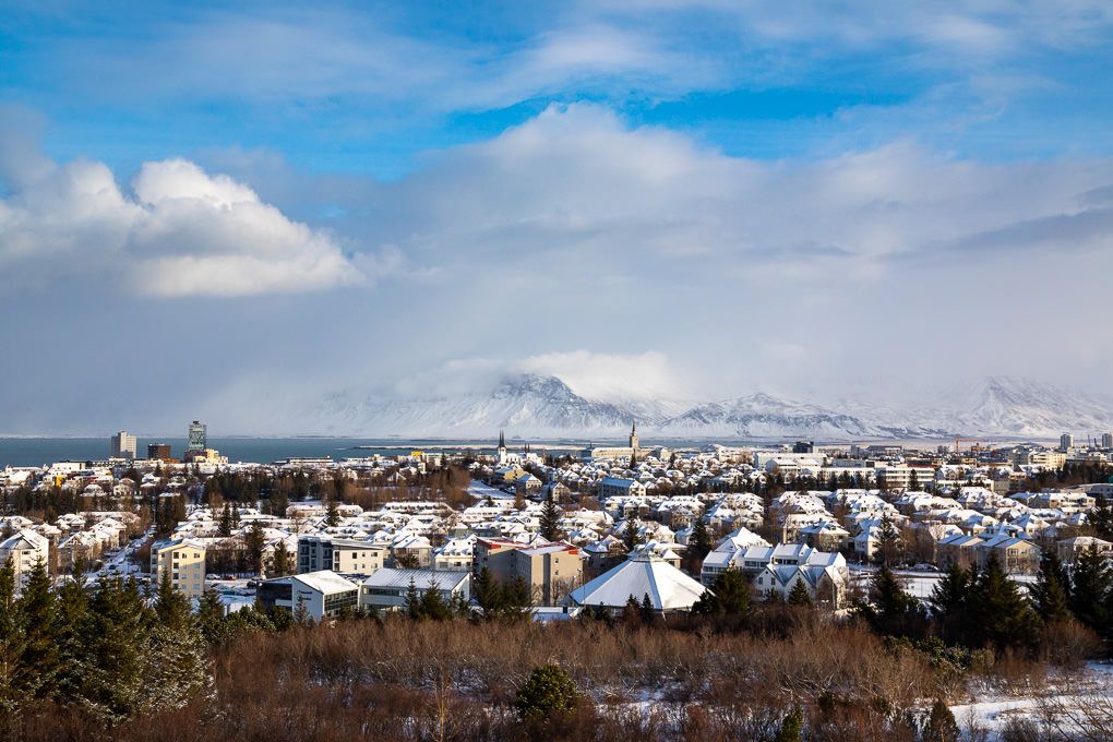 Panoramic cityscape of Reykjavik with snow-covered buildings and mountains under a blue winter sky with scattered clouds.