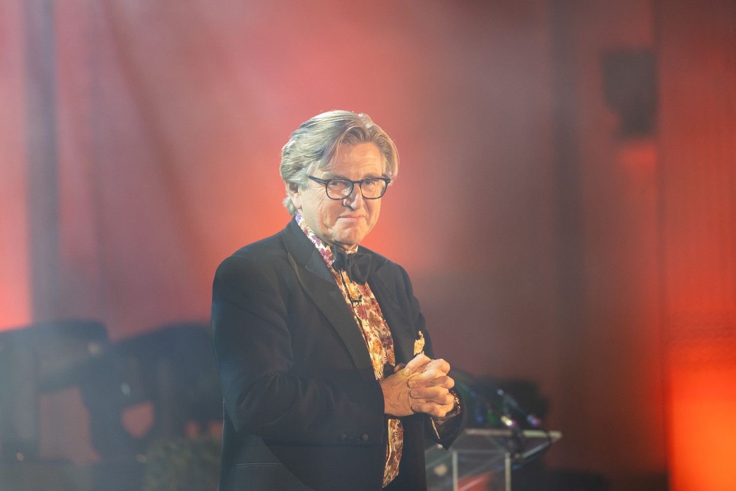 London Event Photography. A male host in glasses and formal attire talks on stage with dramatic red lighting in the background.