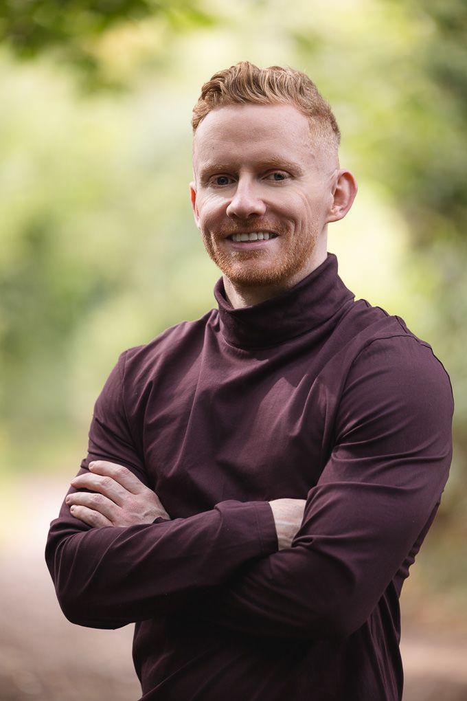 A person with red hair smiling and wearing a brown hoodie poses in different positions outdoors against a blurred background.