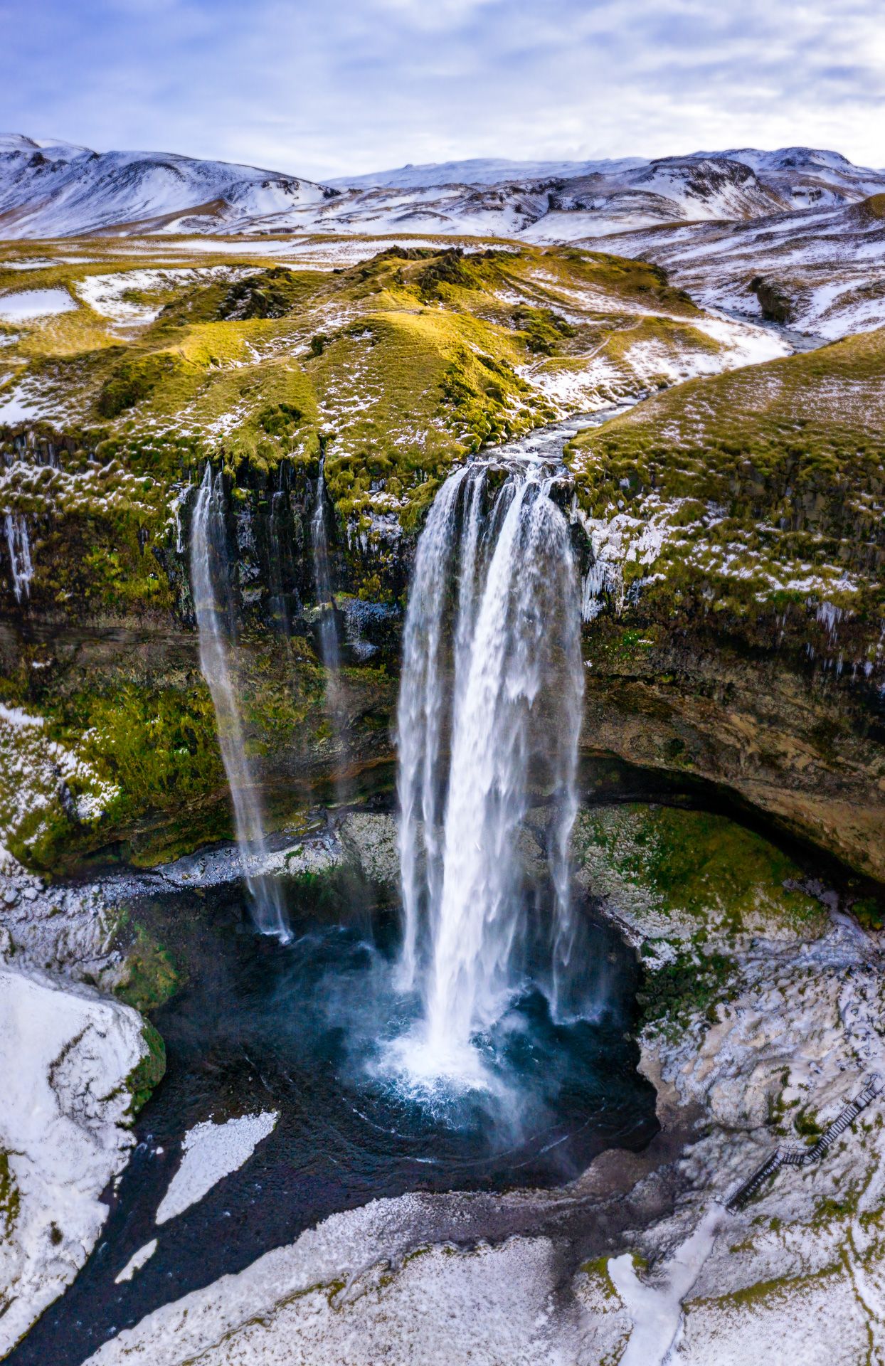 Dramatic aerial view of Seljalandsfoss waterfall in Iceland with snow-capped mountains and green moss-covered cliffs.