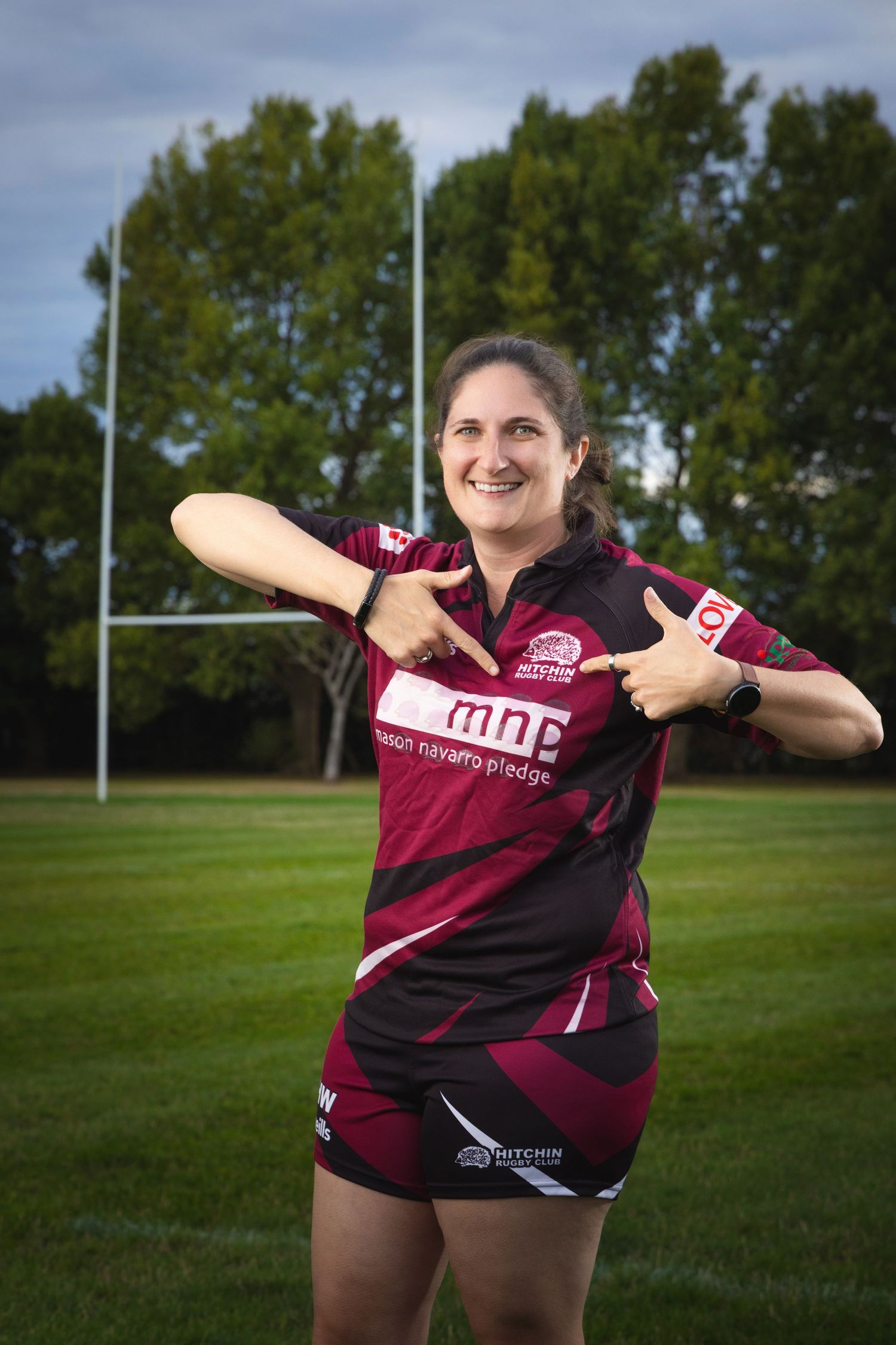 Woman in maroon rugby jersey pointing at MNP logo, smiling on rugby field with goalposts in background.