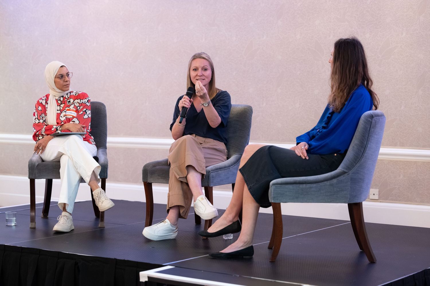 Three women engage in a panel discussion on stage, with one wearing a hijab, at a professional indoor event.