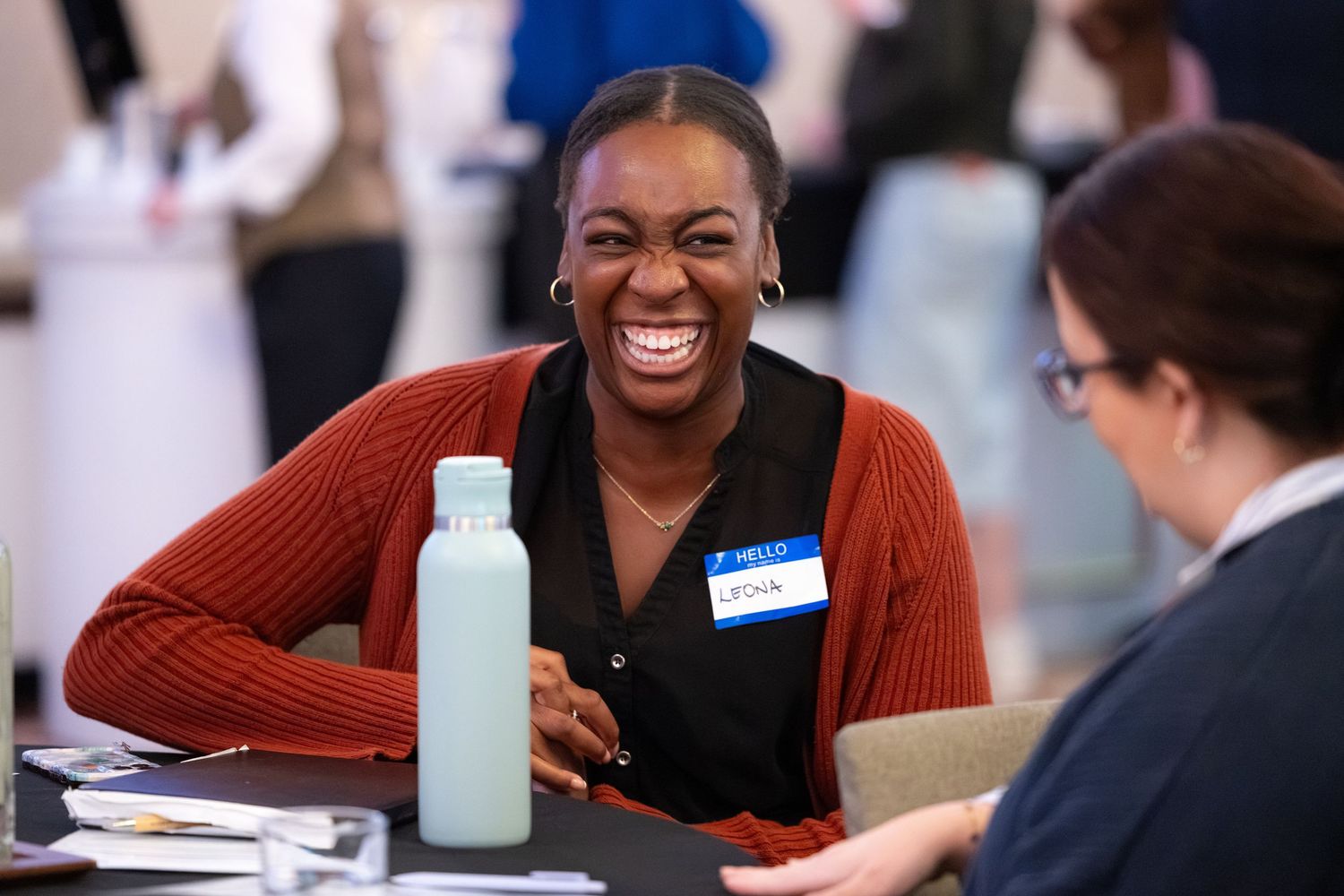 A woman laughs joyfully at a networking event, wearing a name tag and sitting across from another attendee.