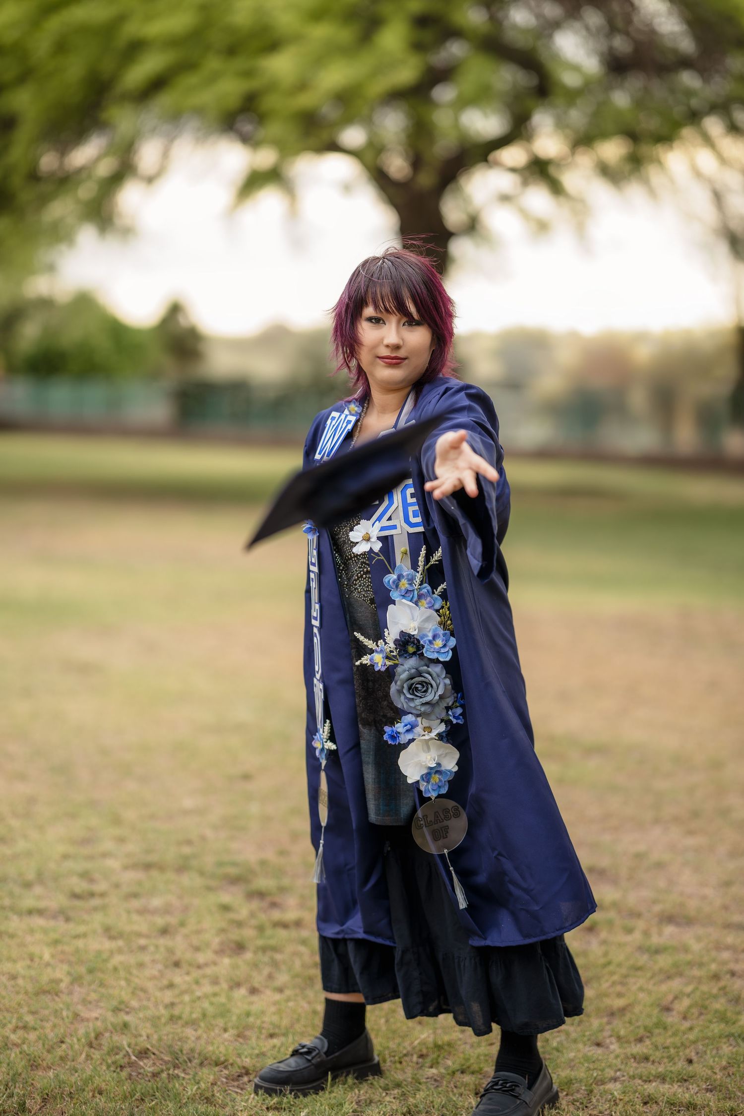 Woman in blue floral embroidered cosplay cape poses outdoors in a park, extending her arm dramatically.