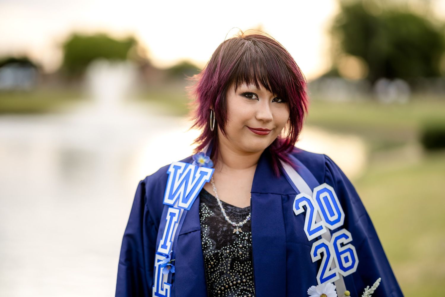 Graduate wearing blue cap and gown with 2026 sash, posing outdoors near a fountain with colorful hair.