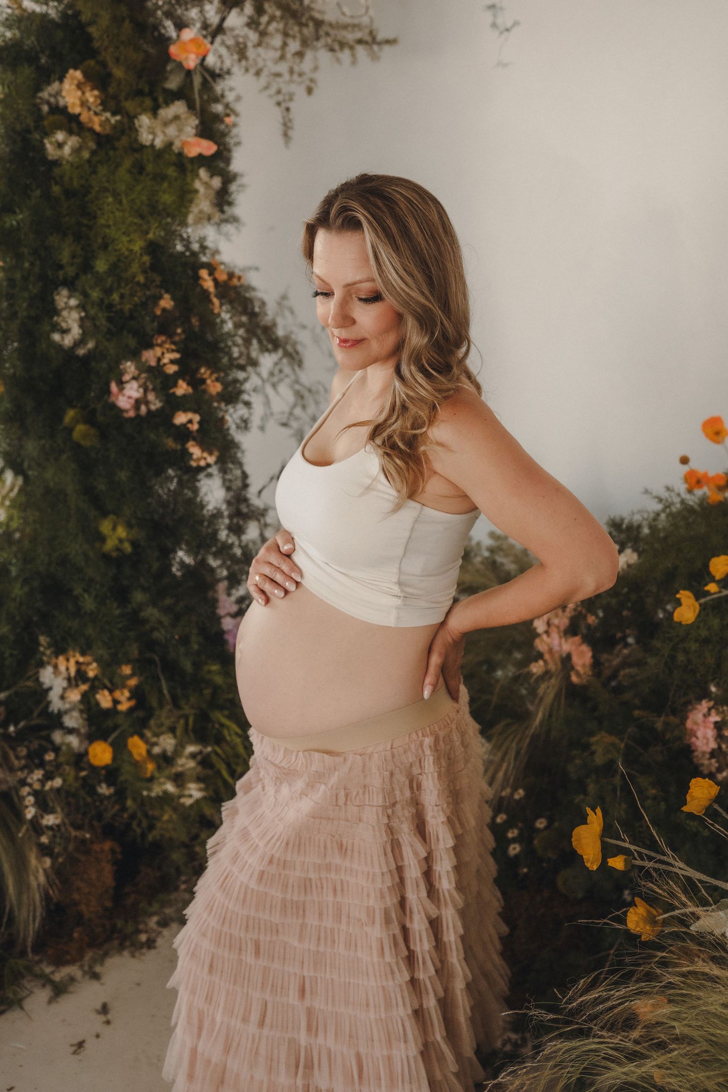 Pregnant woman in white crop top and pink tulle skirt posing among colorful floral arrangement.
