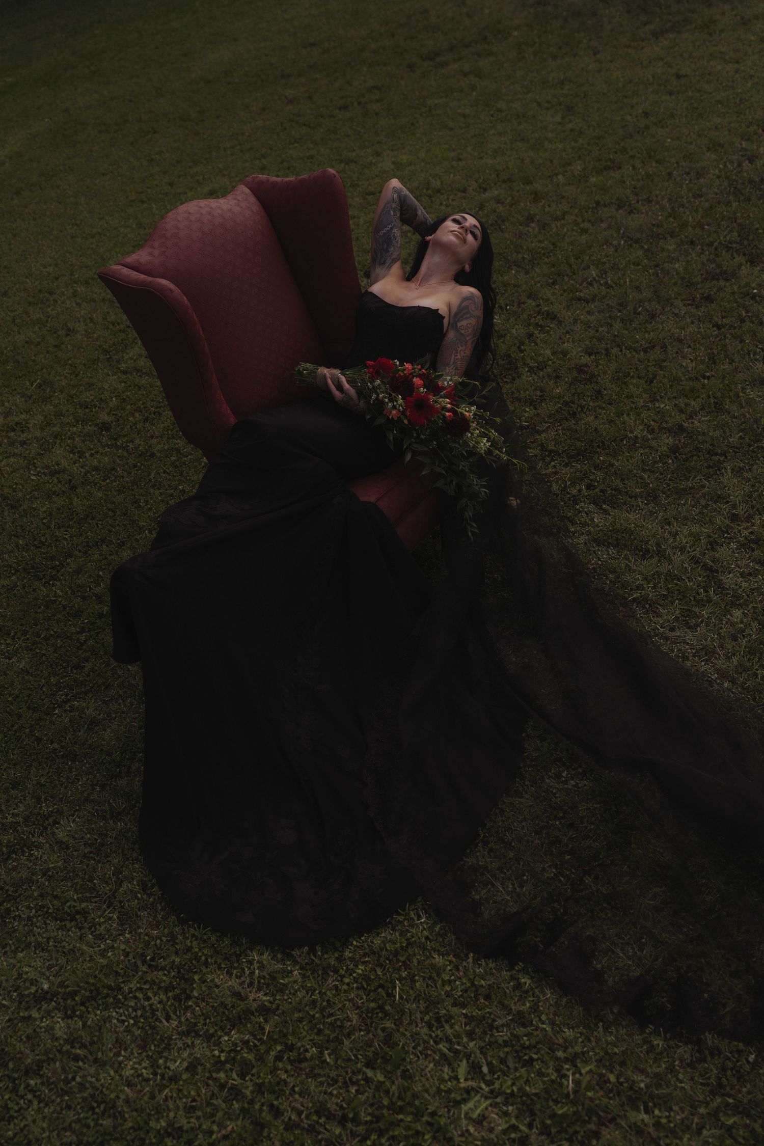 Woman in black gown reclining in red velvet chair on grass holding red flower bouquet.