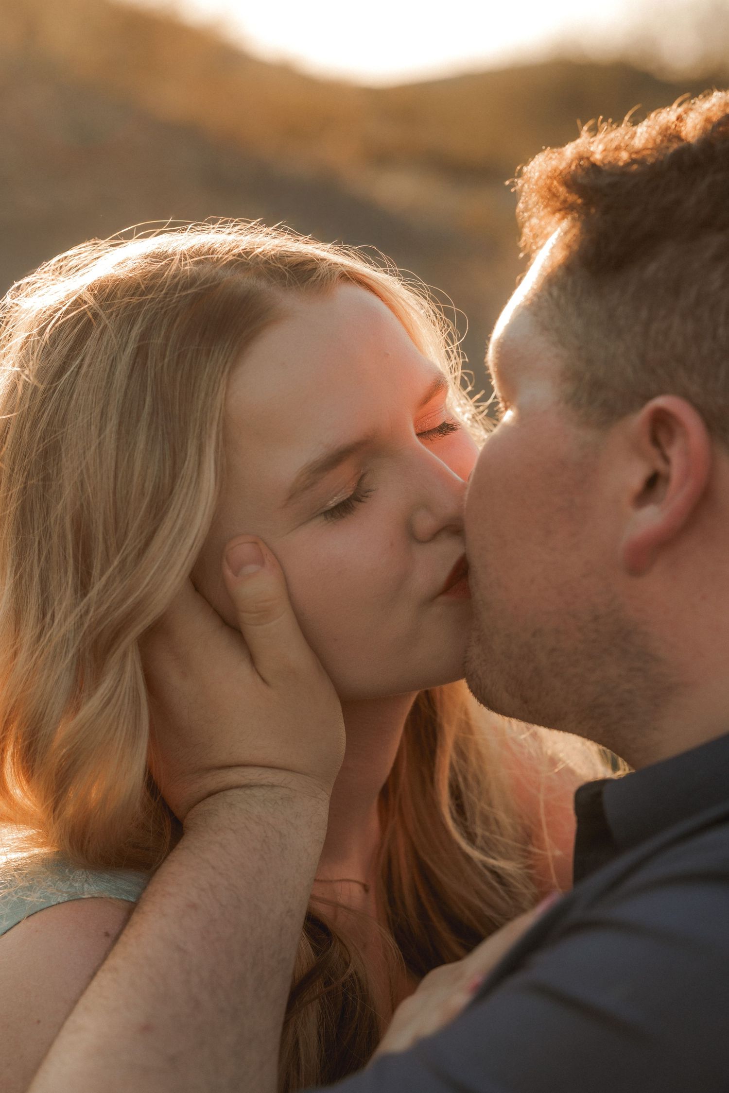 Couple sharing a tender kiss at golden hour with warm sunlight in the background.