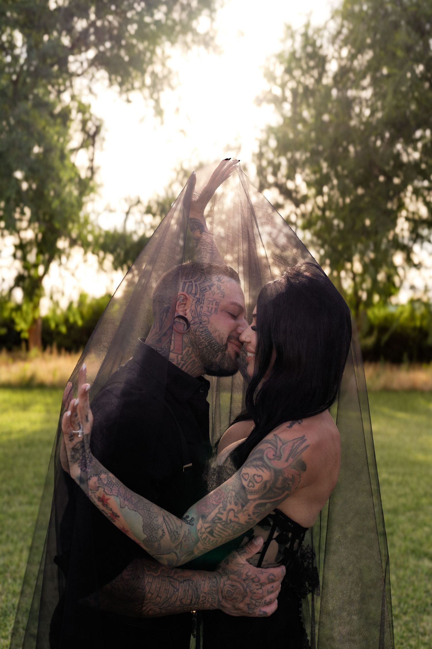 Tattooed couple sharing a romantic kiss under a dark veil in a sunlit garden.