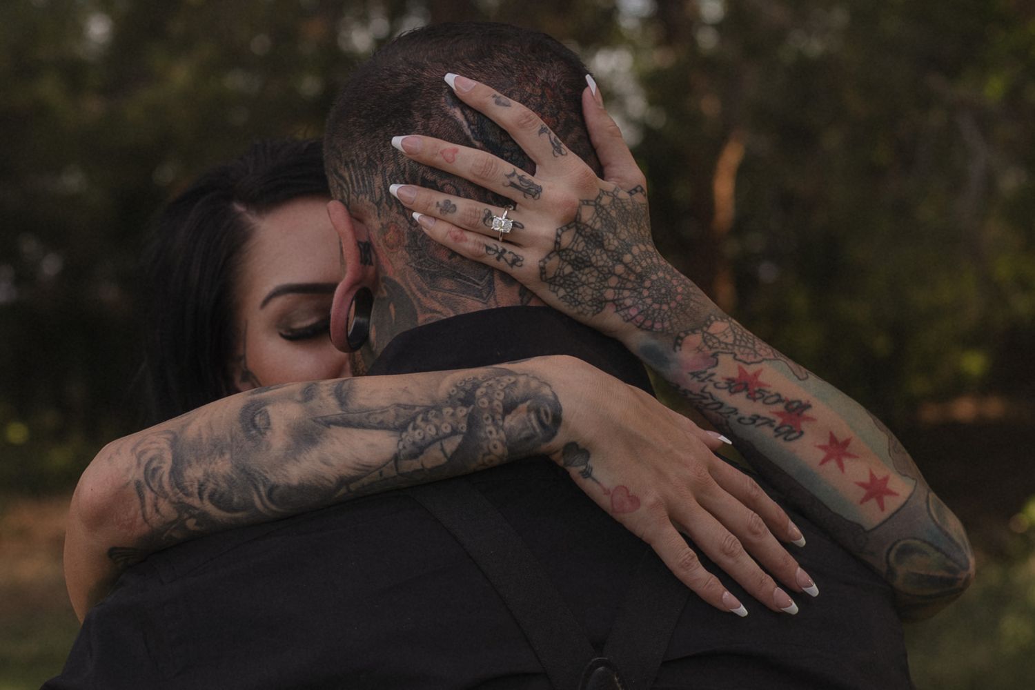Tattooed couple embraces tenderly outdoors, showcasing intricate tattoo artwork on their arms.