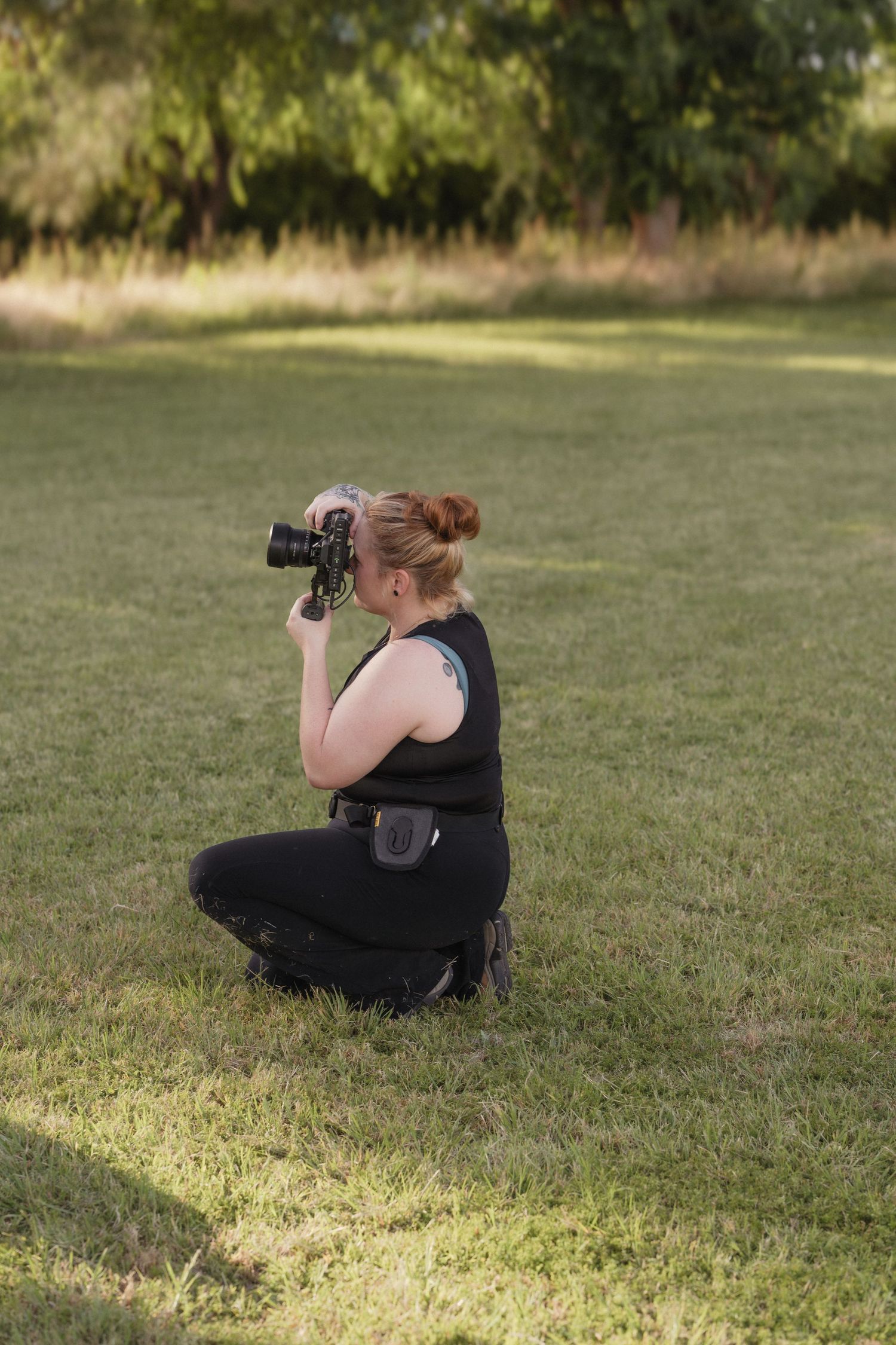 Female photographer crouching on grass in park capturing photos with professional camera.