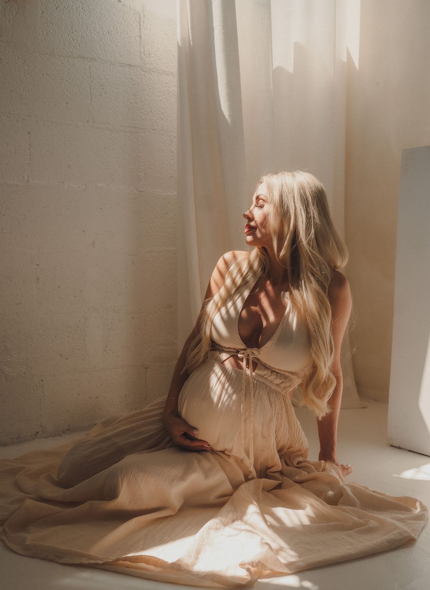 Pregnant blonde woman in a beige dress sitting on the floor in soft natural light.