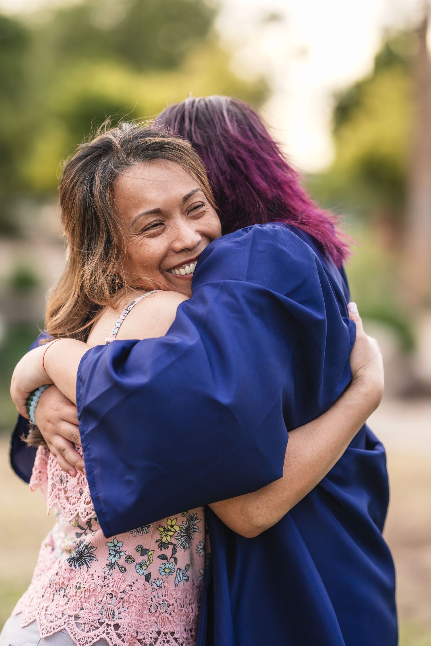 A smiling woman hugs a graduate in a blue cap and gown outdoors during a graduation celebration.