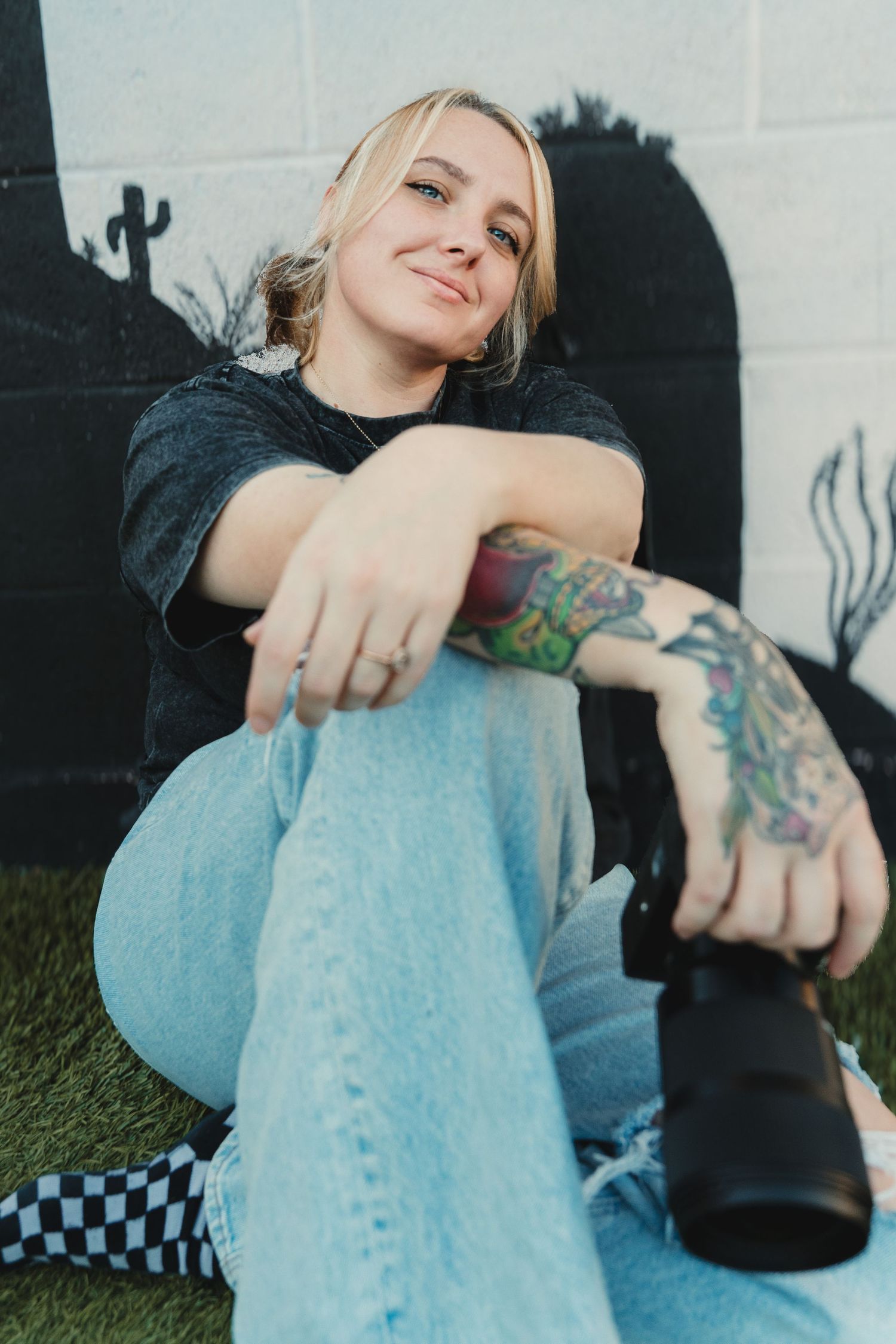 Smiling blonde woman with colorful arm tattoos sits on grass holding a camera.