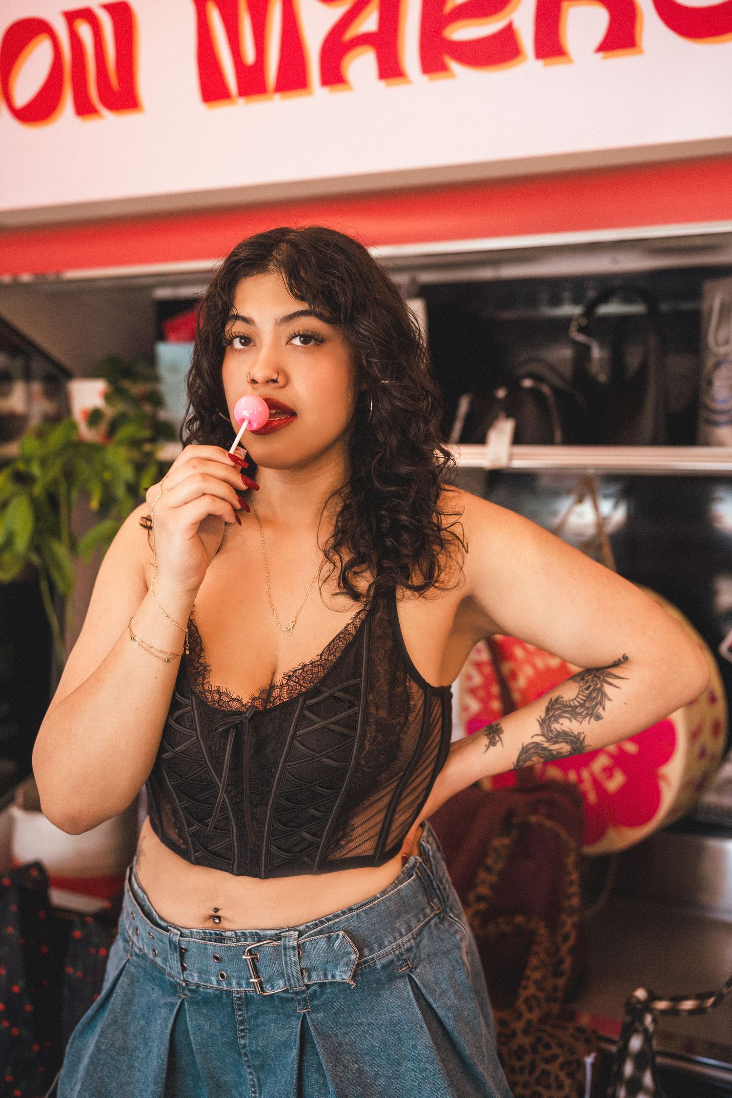 Woman in black lace crop top and denim skirt eating a pink lollipop near a food truck.