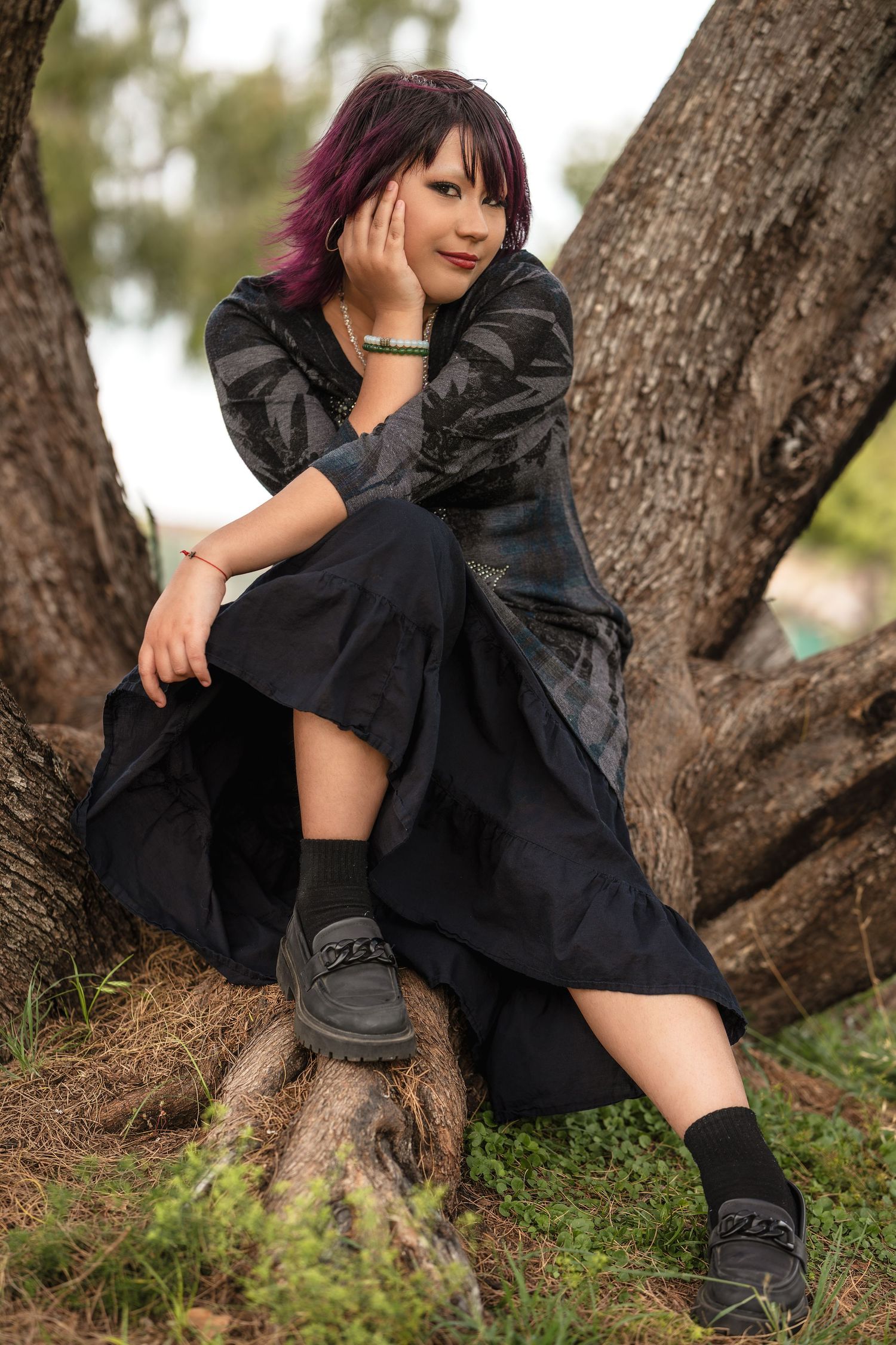 Young woman with purple hair in black dress sits against tree roots outdoors.