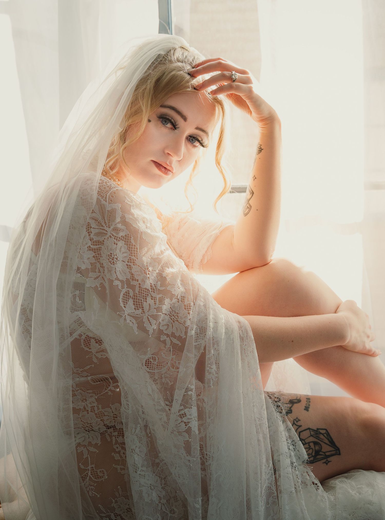 Blonde bride in sheer lace robe and veil sits gracefully by a sunlit window with tattoos visible.
