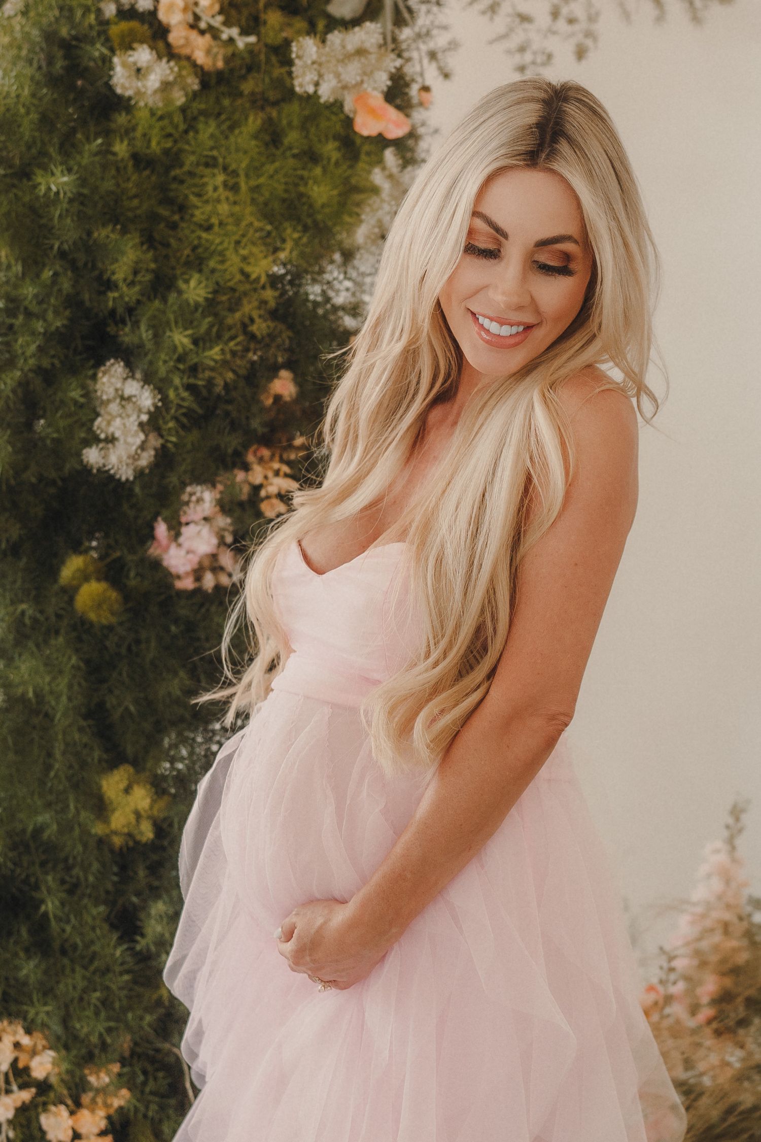 Smiling pregnant woman in pink tulle gown with long blonde hair posing in front of floral wall.